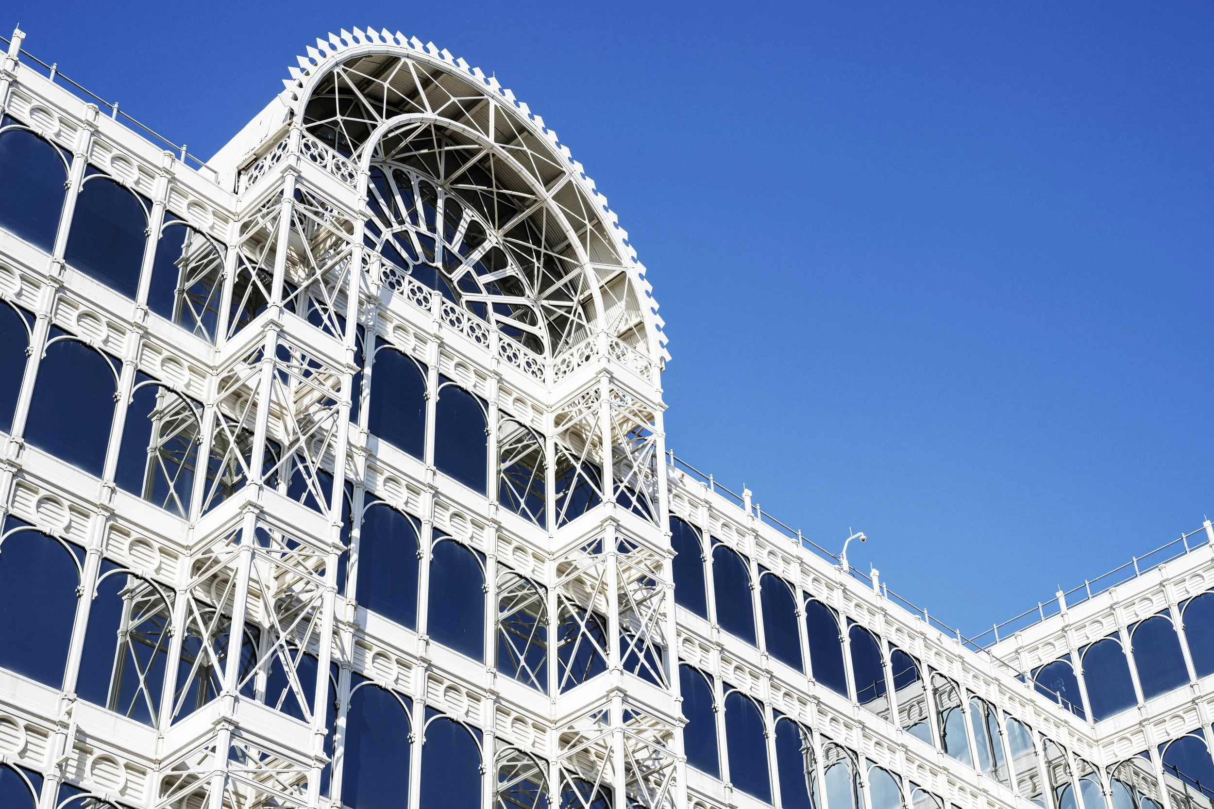 A white, ornate, multi-level building with a large arch at the top, featuring many decorative metal arches and large windows, set against a clear blue sky.