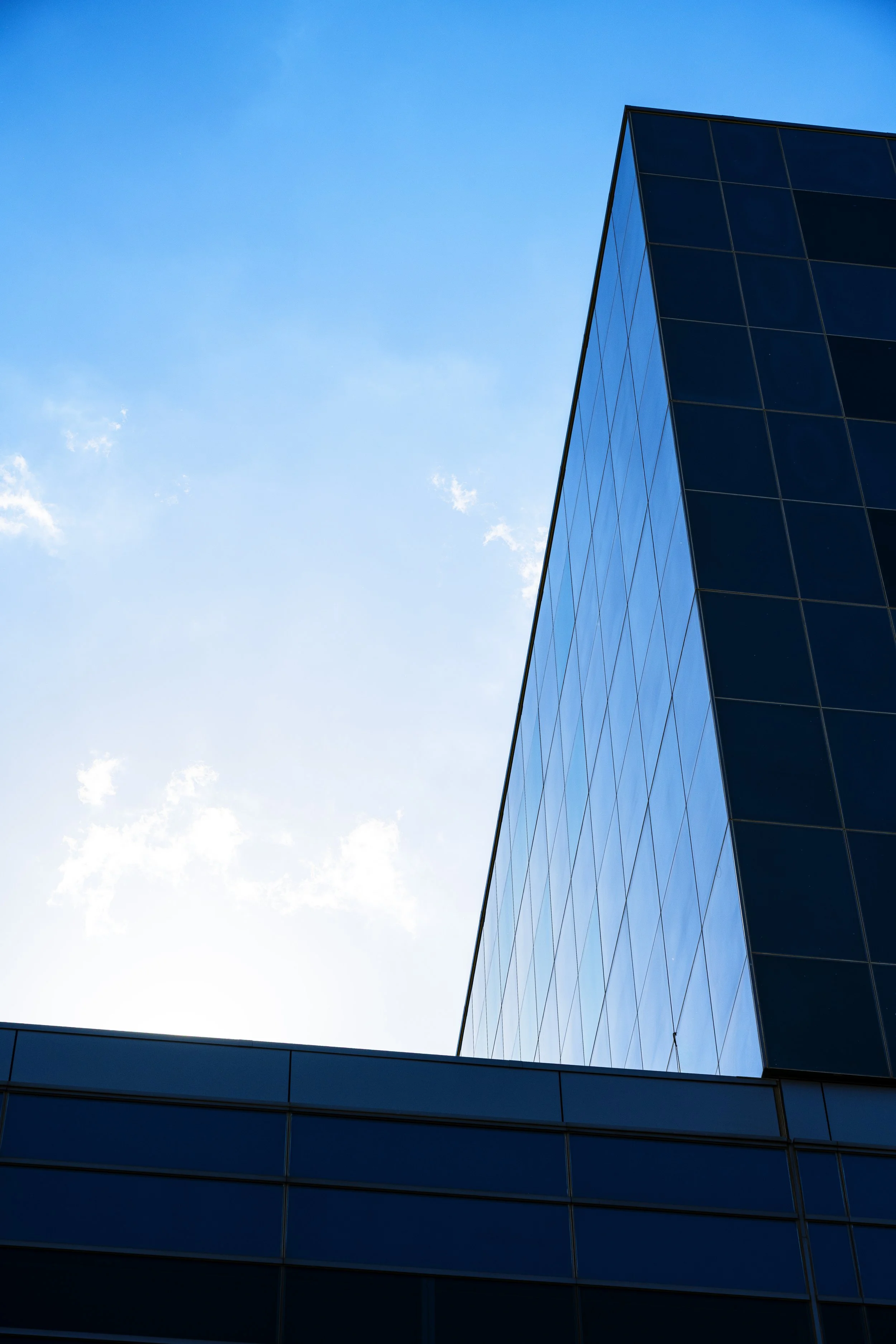 The photo features a modern glass building with a sleek black facade in the lower part of the image and a clear blue sky with a few wispy clouds in the background. The building's reflective glass panels mirror the sky.