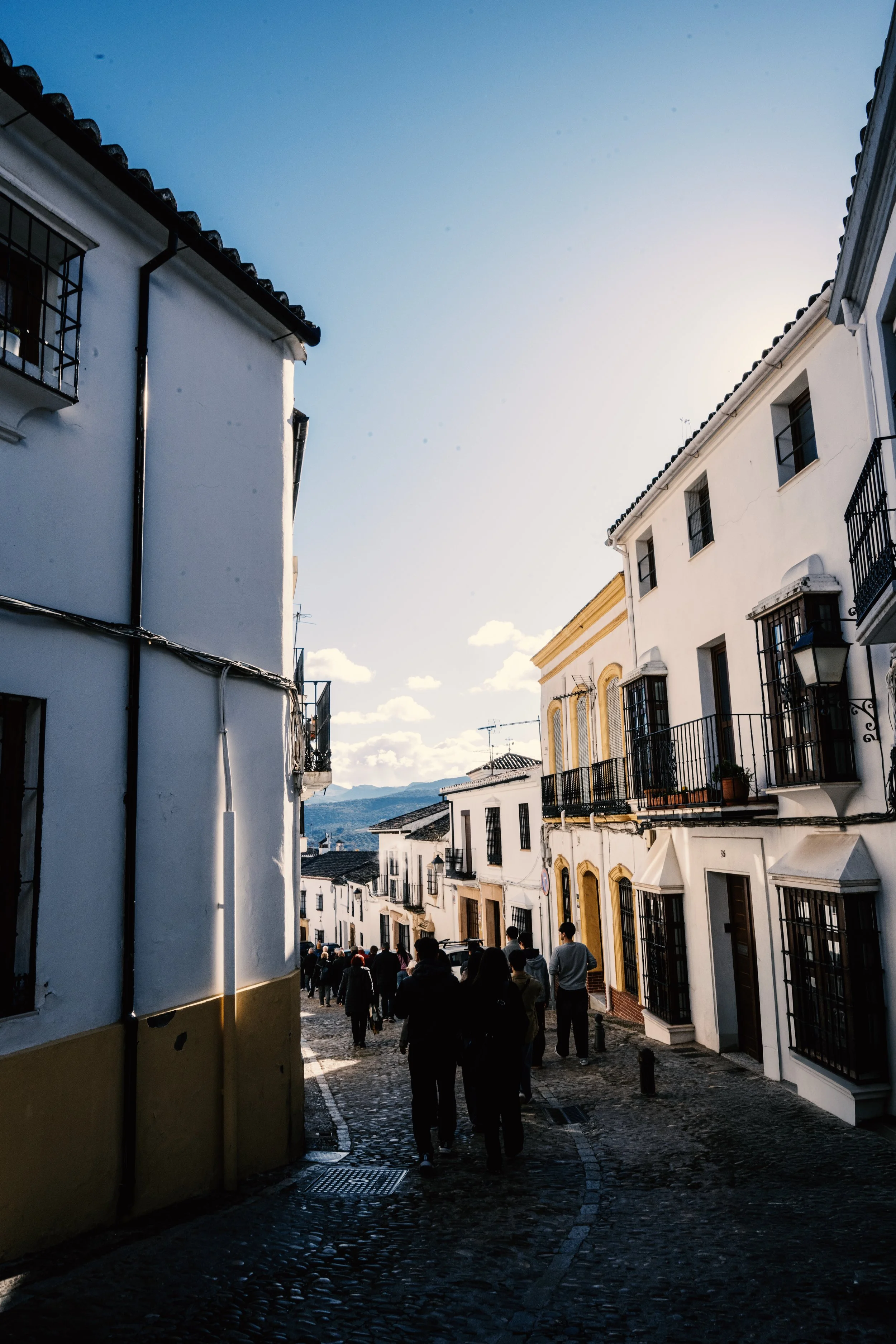 People walking down a narrow cobblestone street between white buildings with balconies, under a clear blue sky with some clouds and distant mountains.