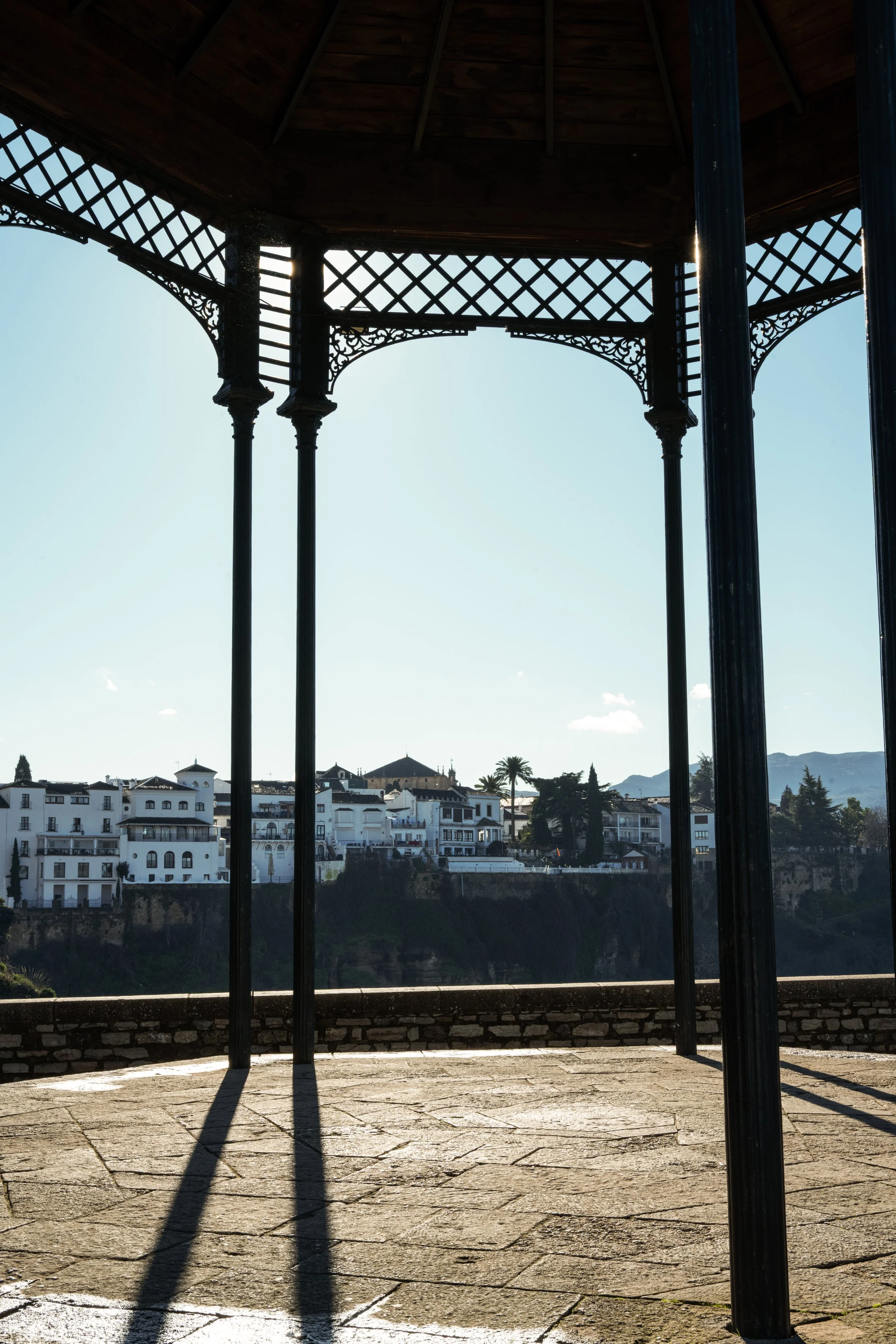 An ornate, open-air gazebo with decorative metalwork, supported by tall, dark metal columns. In the background, white buildings with red-tiled roofs and a landscape of trees and distant mountains are visible under a clear, sunny sky.