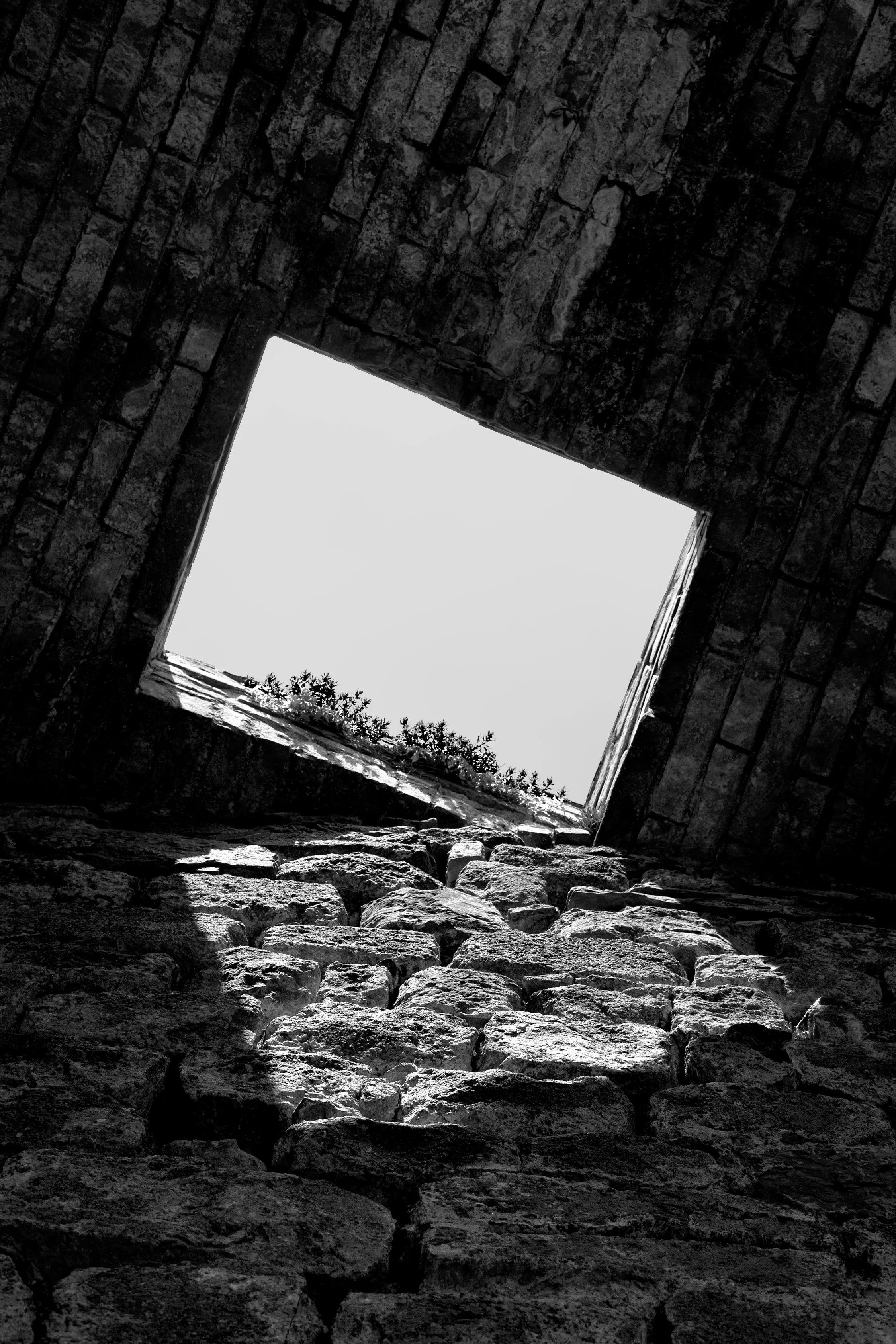 Looking up through a stone well with rough stones at the bottom and brick walls, open to the sky with some trees visible.