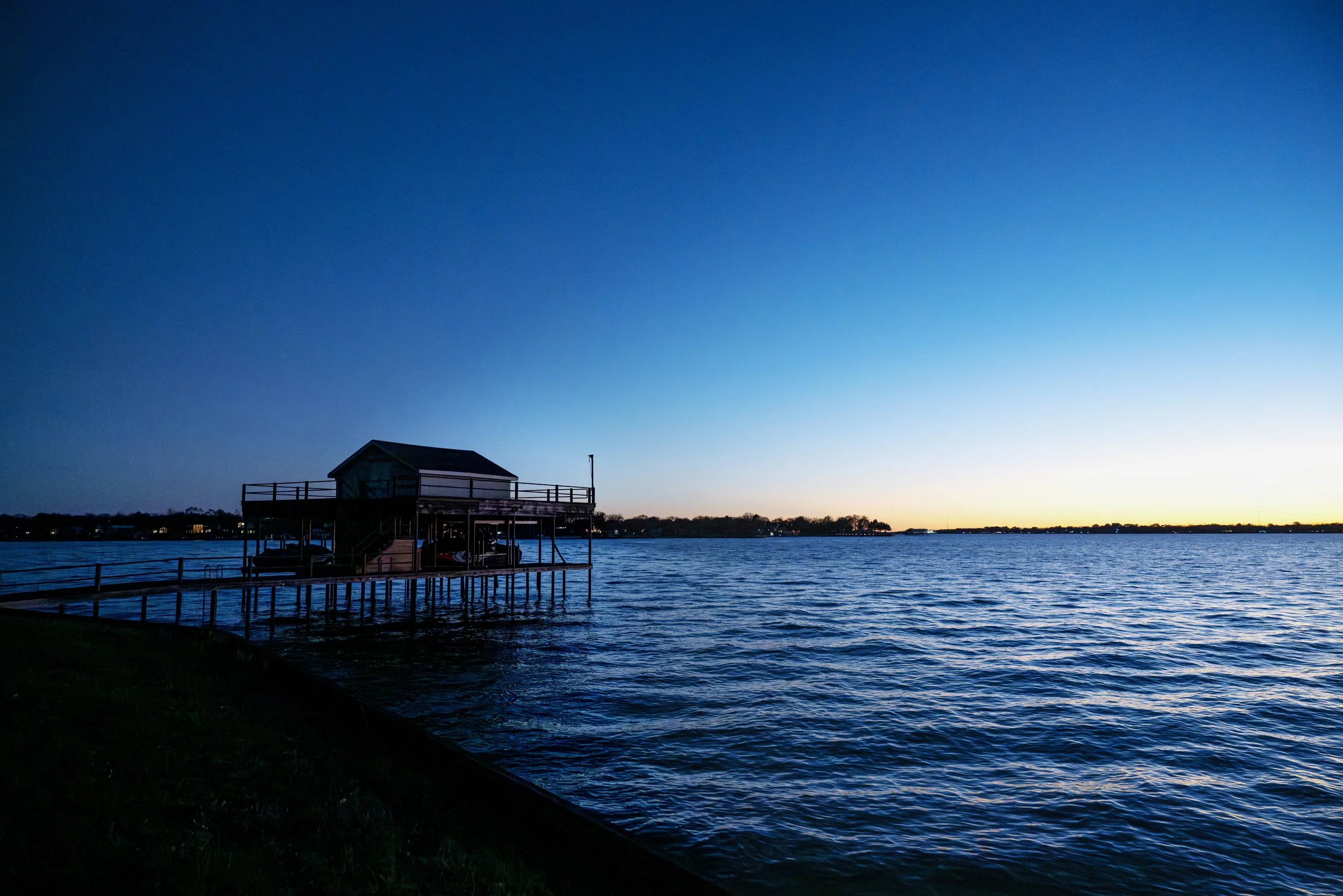 A waterside scene at dusk featuring a small pier with a wooden house on stilts over the water. The sky transitions from sunset colors to deep blue, and the water reflects the sky's hues, with trees and houses in the distant background.