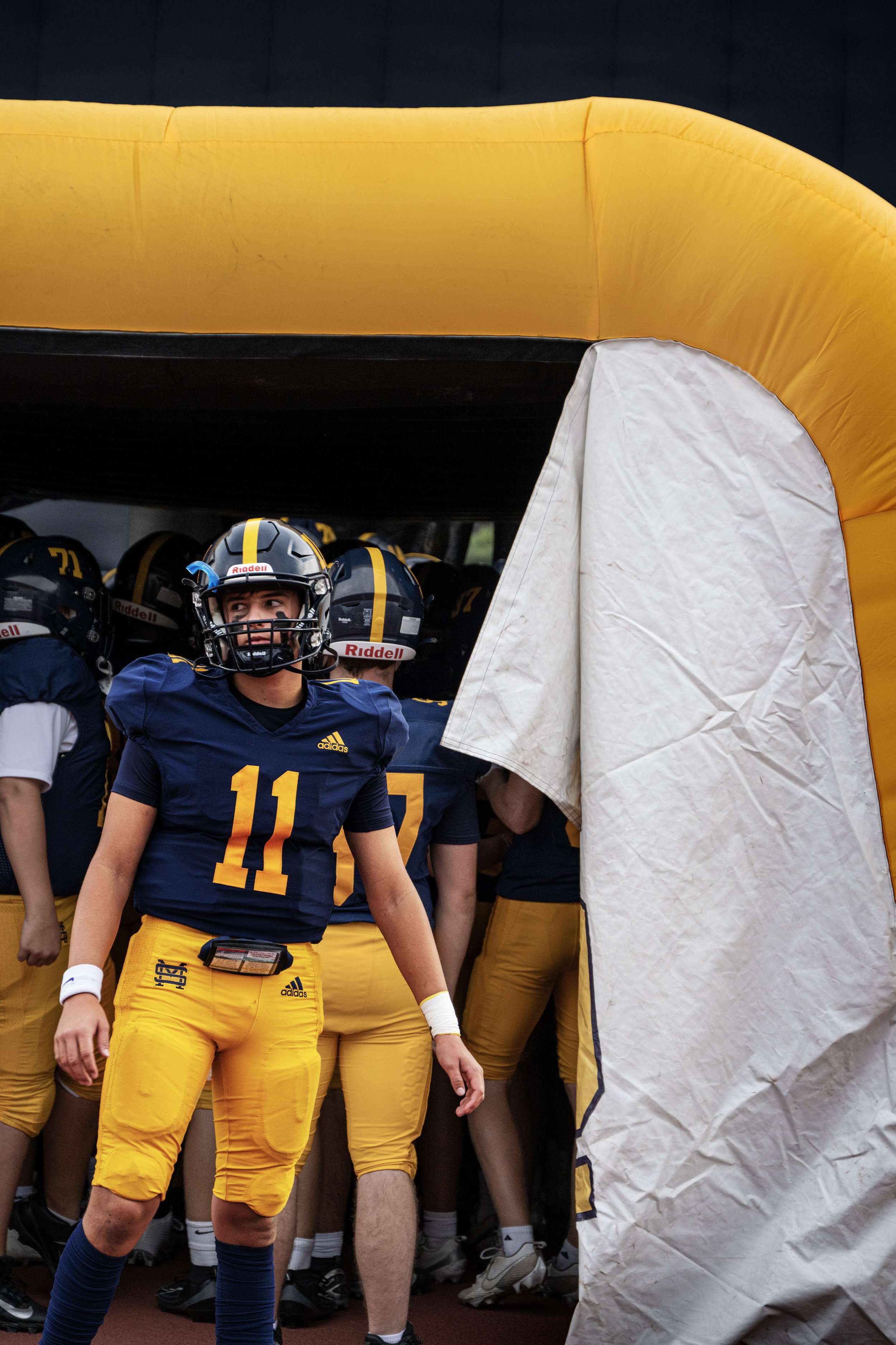 American football team coming out of tunnel onto field, player wearing jersey number 11 in the front, other team members in the background, under a yellow inflatable tunnel.