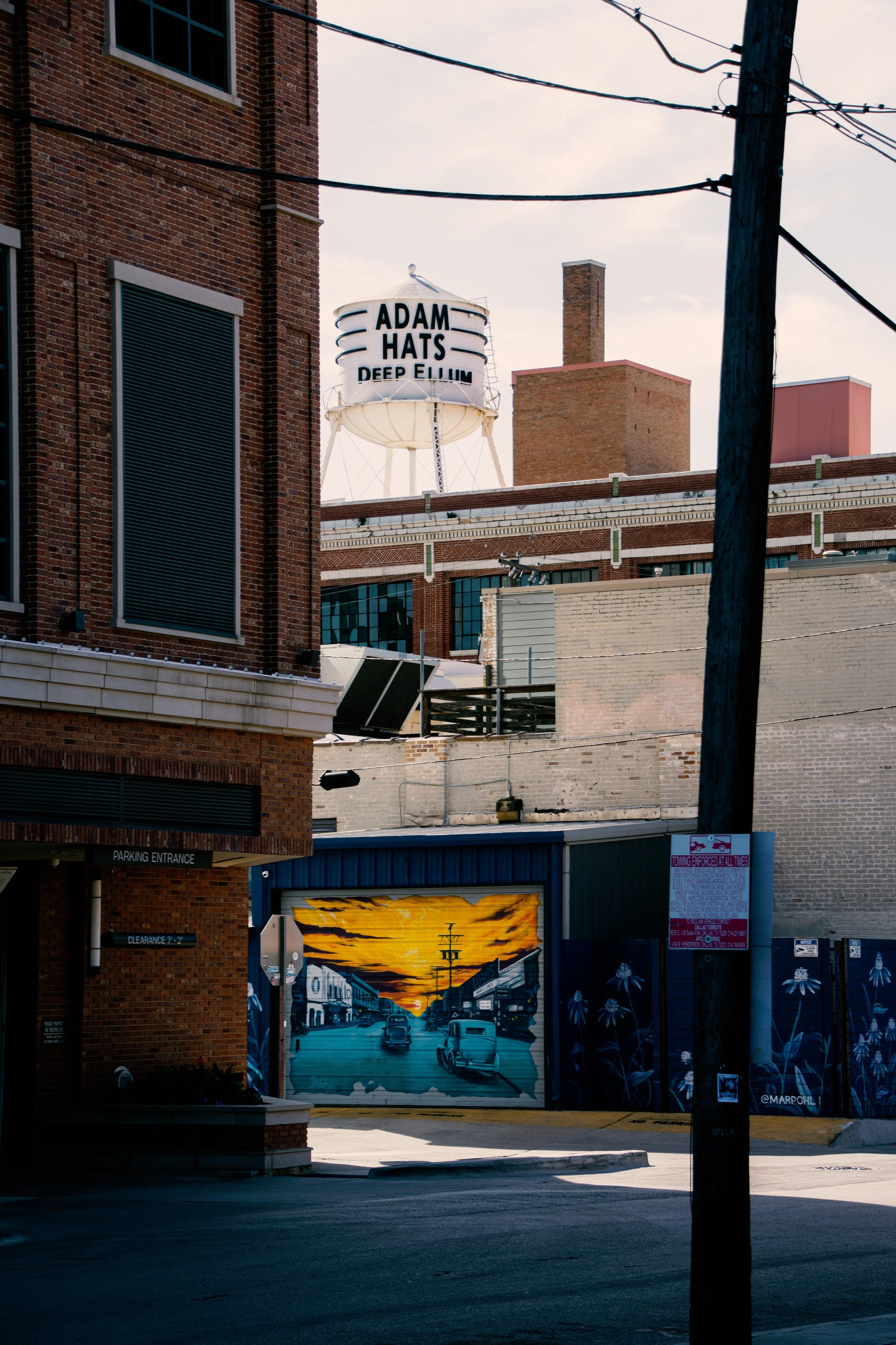 Photograph of a city street scene with brick buildings, utility pole, and a mural of a sunset over a train station painted on a garage door.