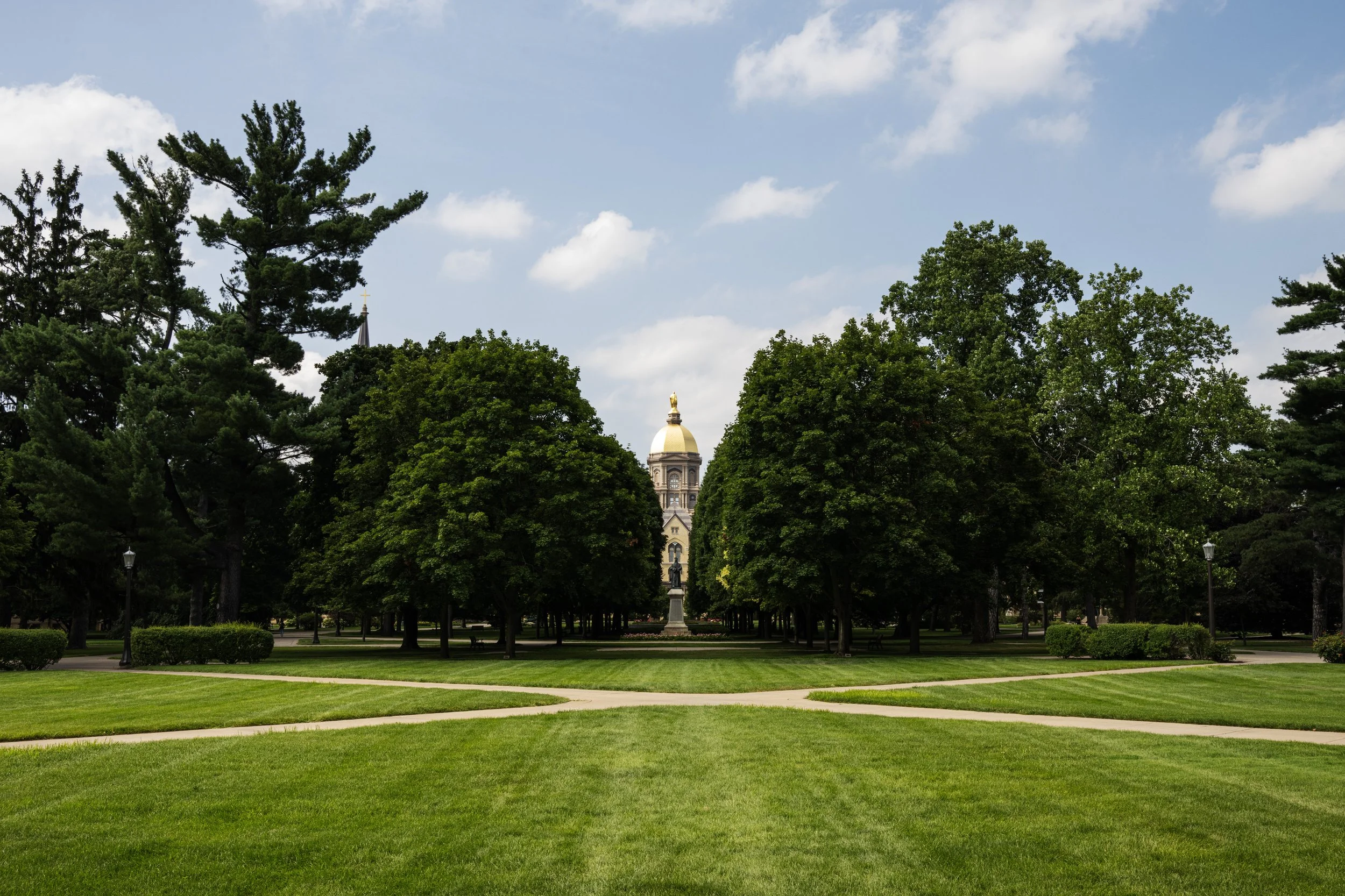 A park with green grass, pathways, tall trees, and a historic building with a gold dome in the background.