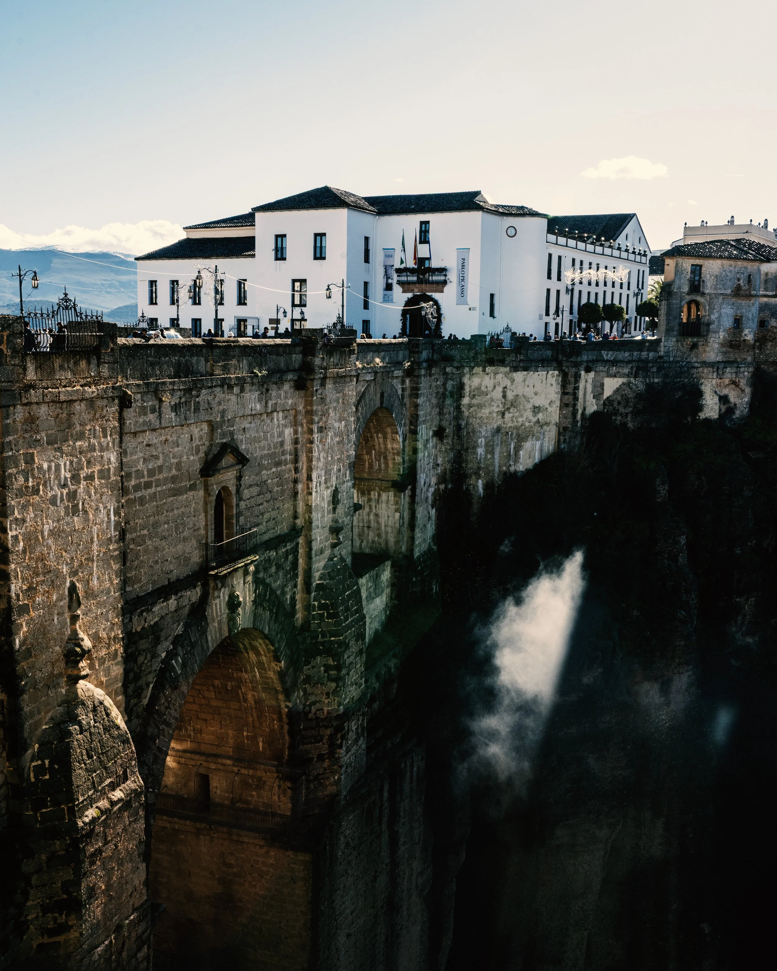 A historic stone bridge with high arches over a river, with a white building with black windows on top, and a mountain range in the background under a clear sky.