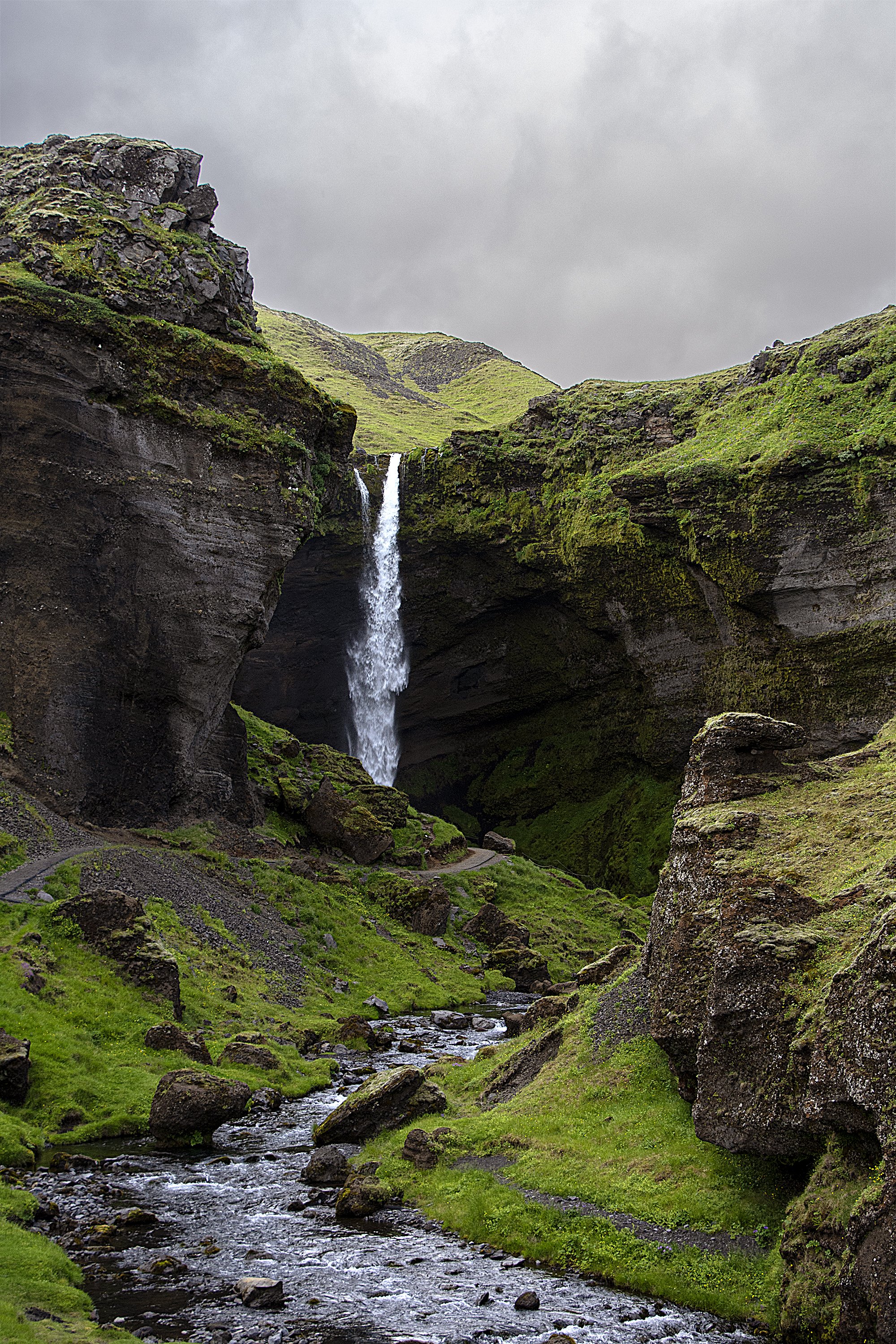 A small waterfall flowing into a stream in a green, moss-covered canyon with rocky cliffs and cloudy sky above.