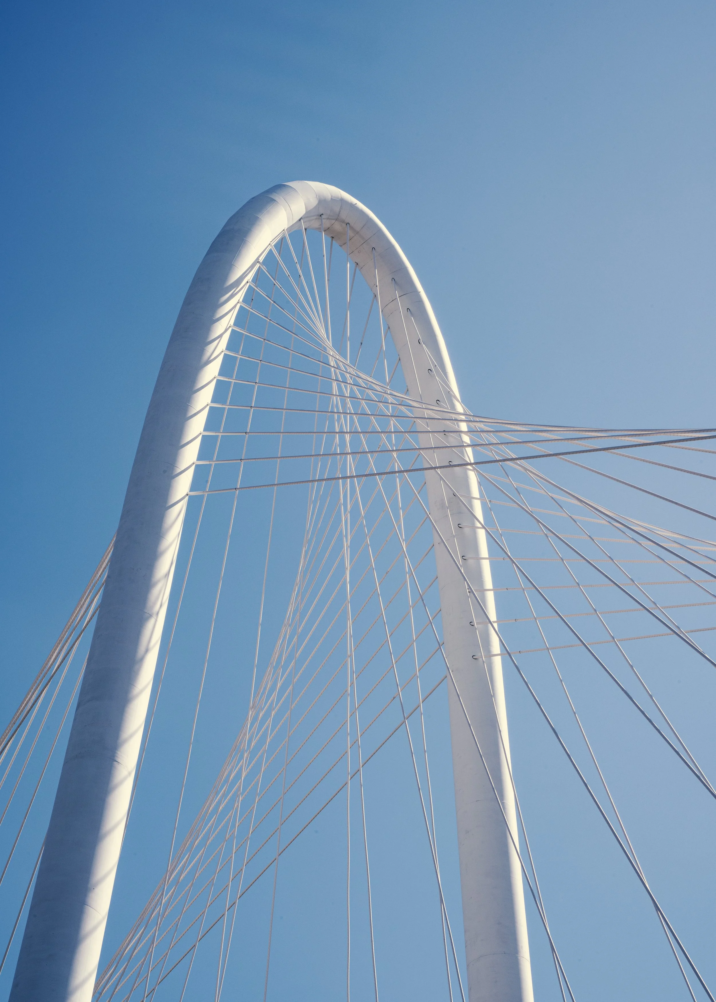 A modern white architectural structure with an arch and radiating cables set against a clear blue sky.