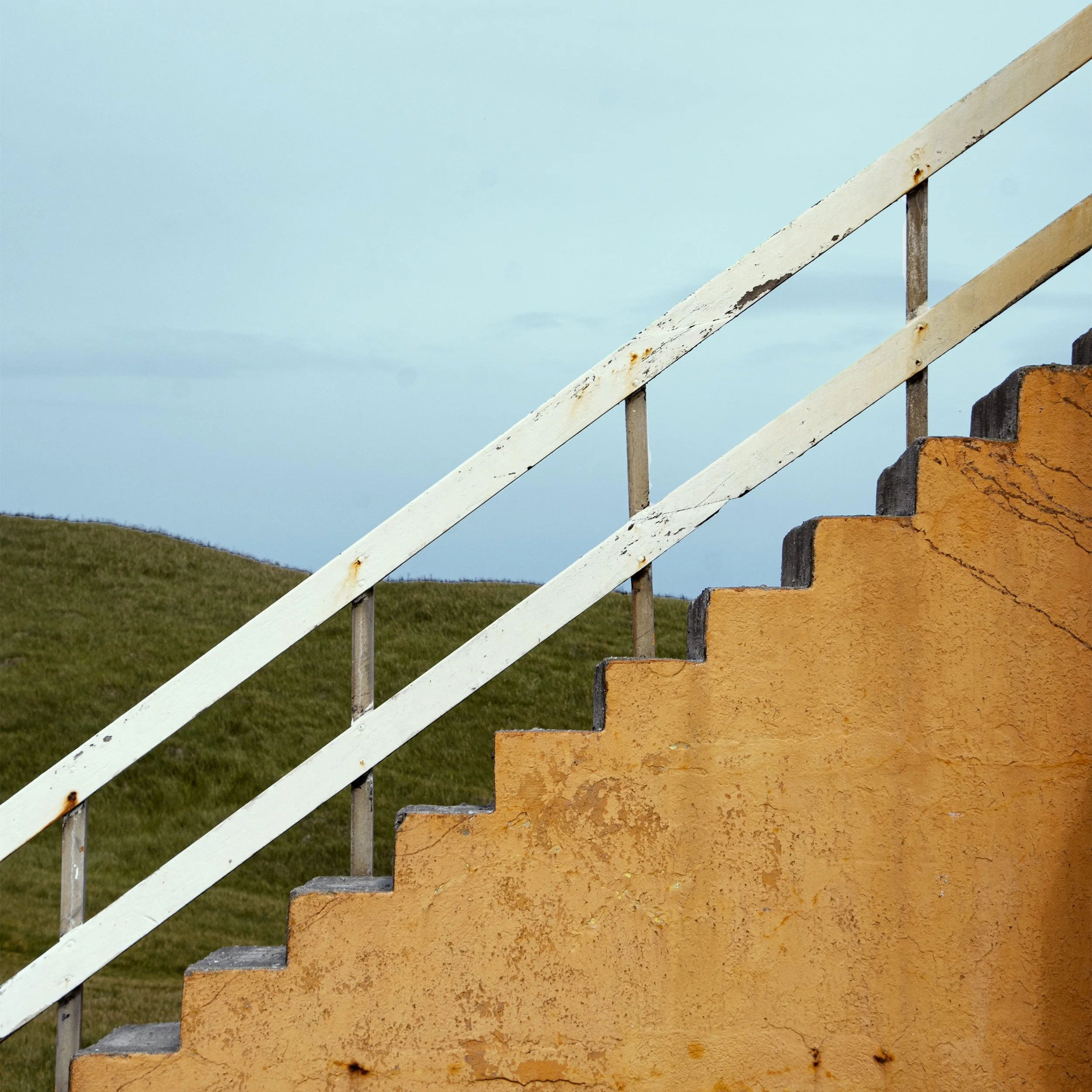 Close-up of a weathered orange concrete staircase with a white metal railing, against a background of a grassy landscape and a cloudy sky.