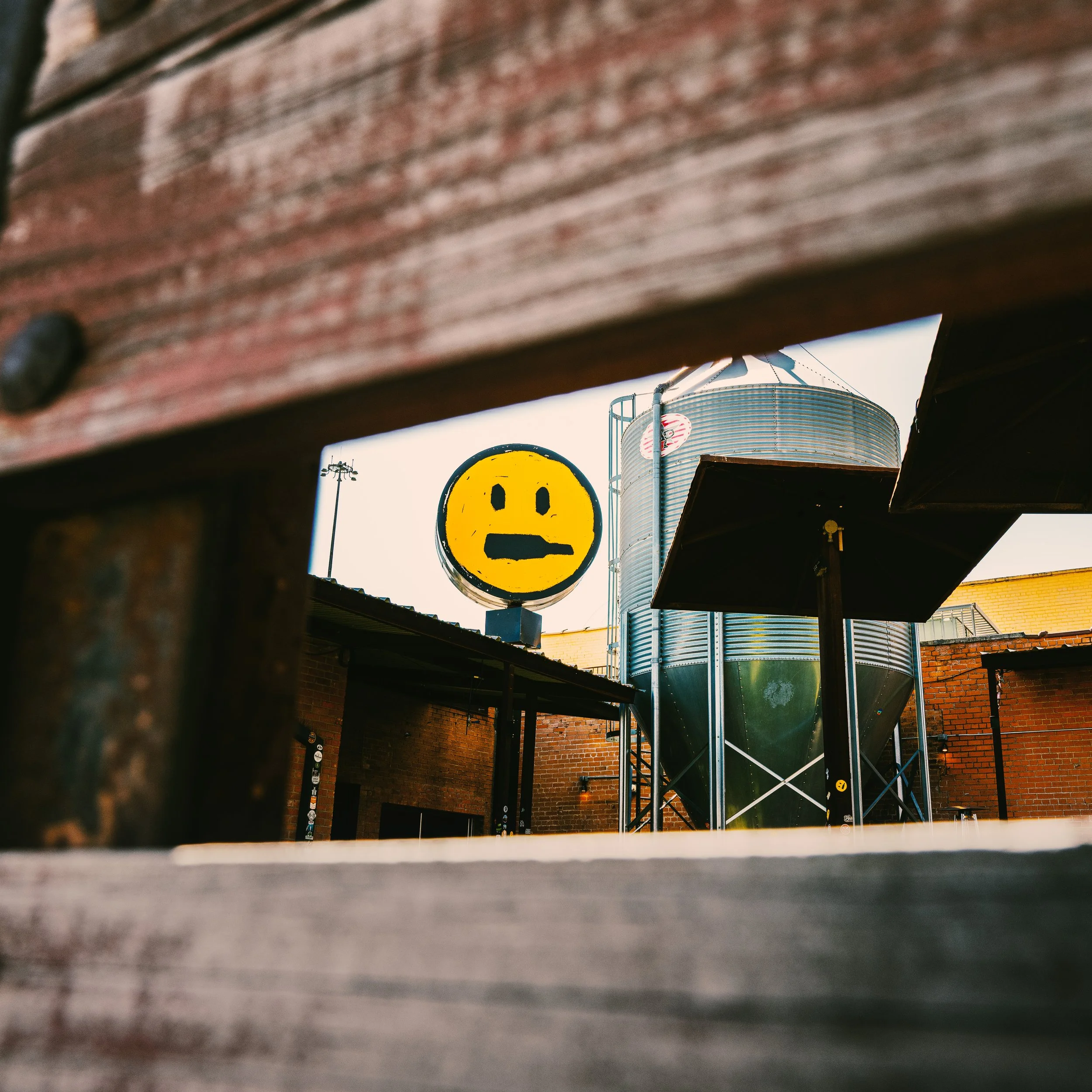 A shot taken from underneath a wooden surface, revealing a water tank with a ladder in the background and a smiley face sign, with power lines and buildings in the distance.