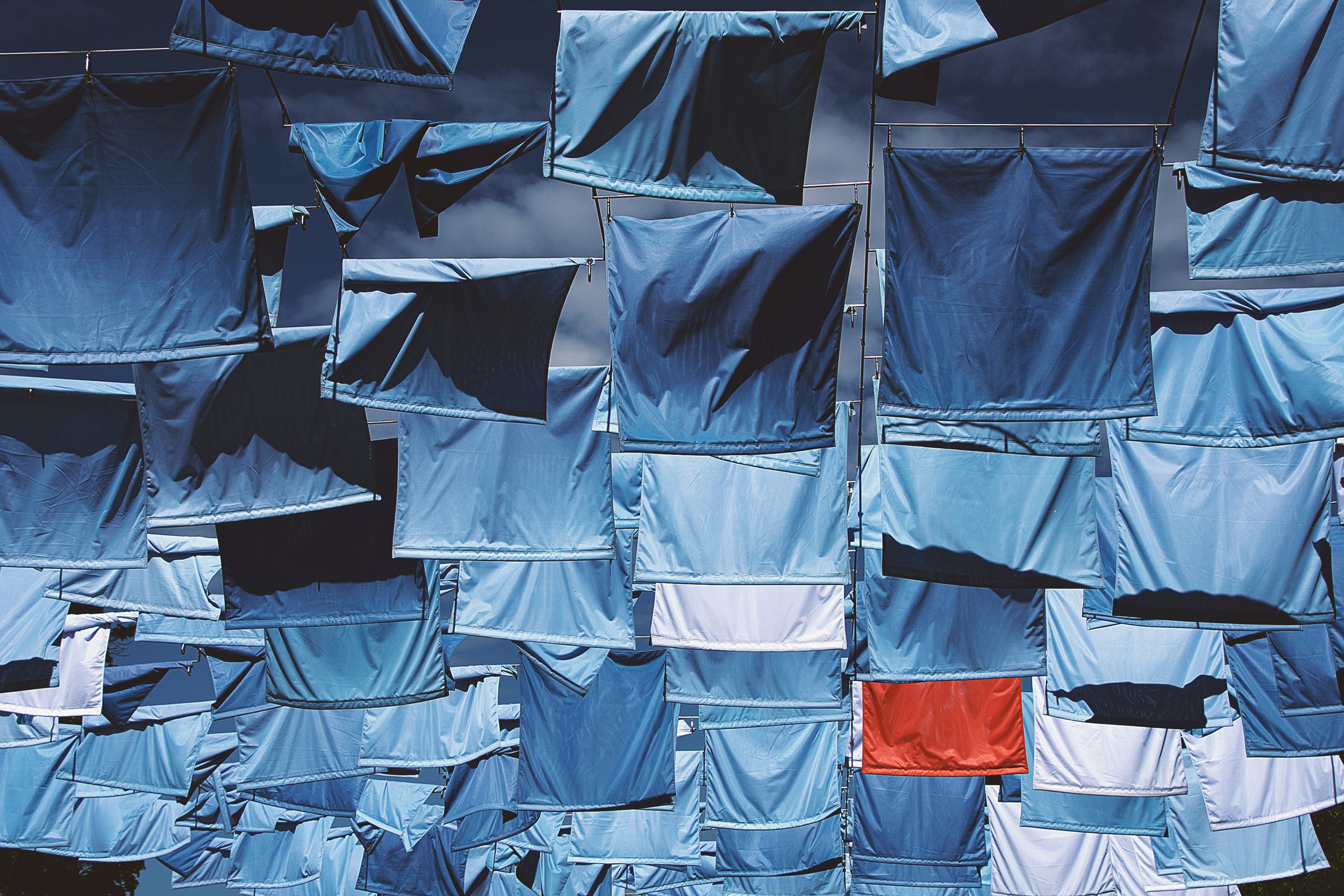 A structure made of numerous blue and white fabric pieces hanging and attached to wires against a dark sky, with a small section of a red piece at the bottom.