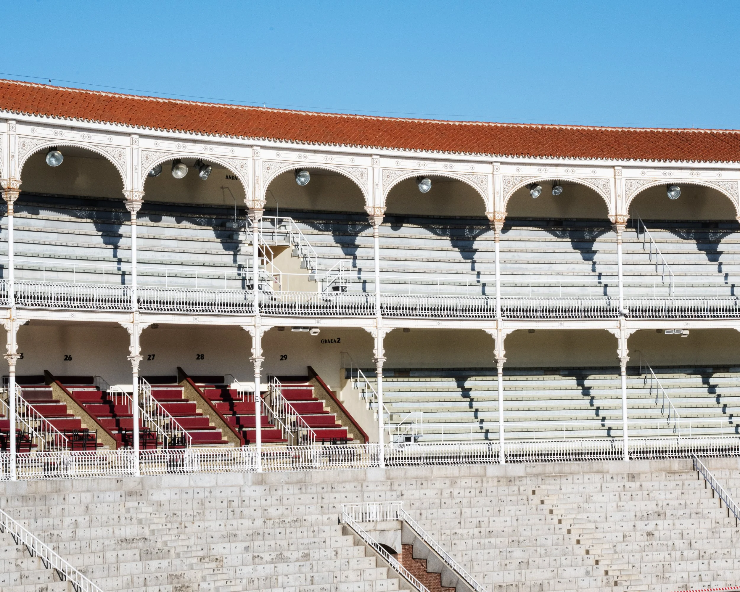Empty tiered stadium with red and gray seating and white decorative arches against a clear blue sky.