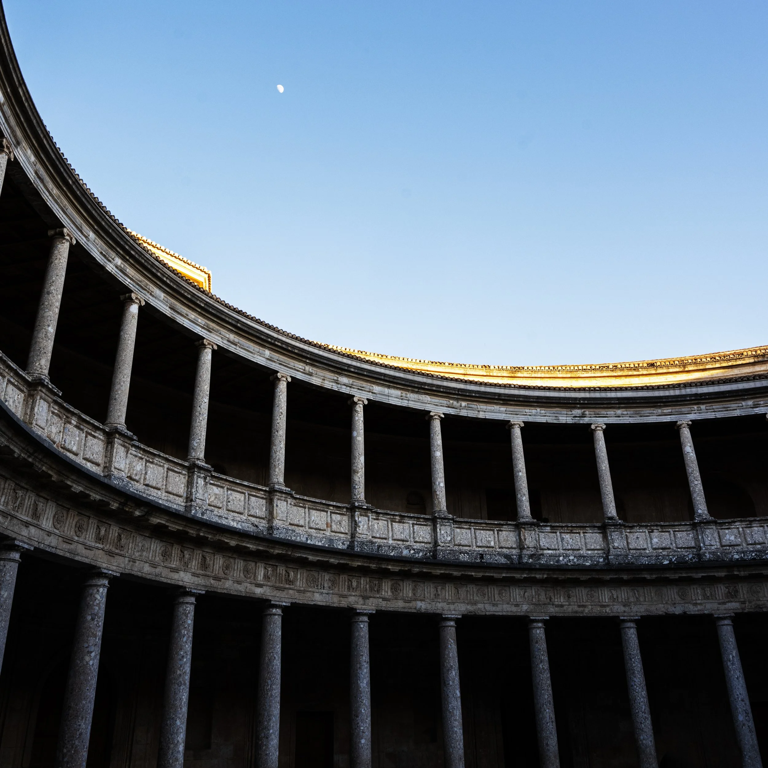 An ancient Roman or Greek colonnade with evenly spaced stone columns supporting a curved entablature, under a clear blue sky with a visible moon.