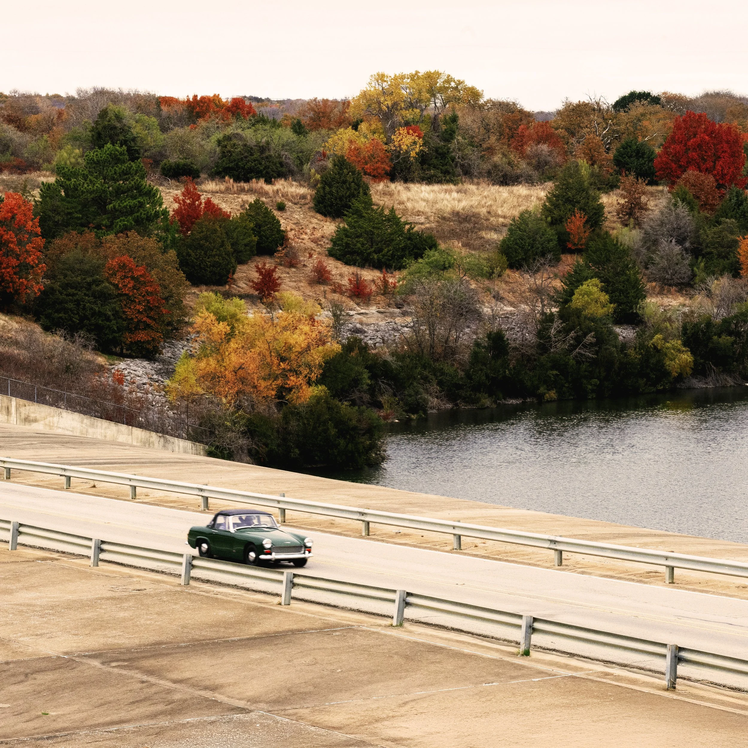A vintage dark green convertible car driving on a paved road beside a body of water, with a landscape of trees in fall colors on a hill in the background.