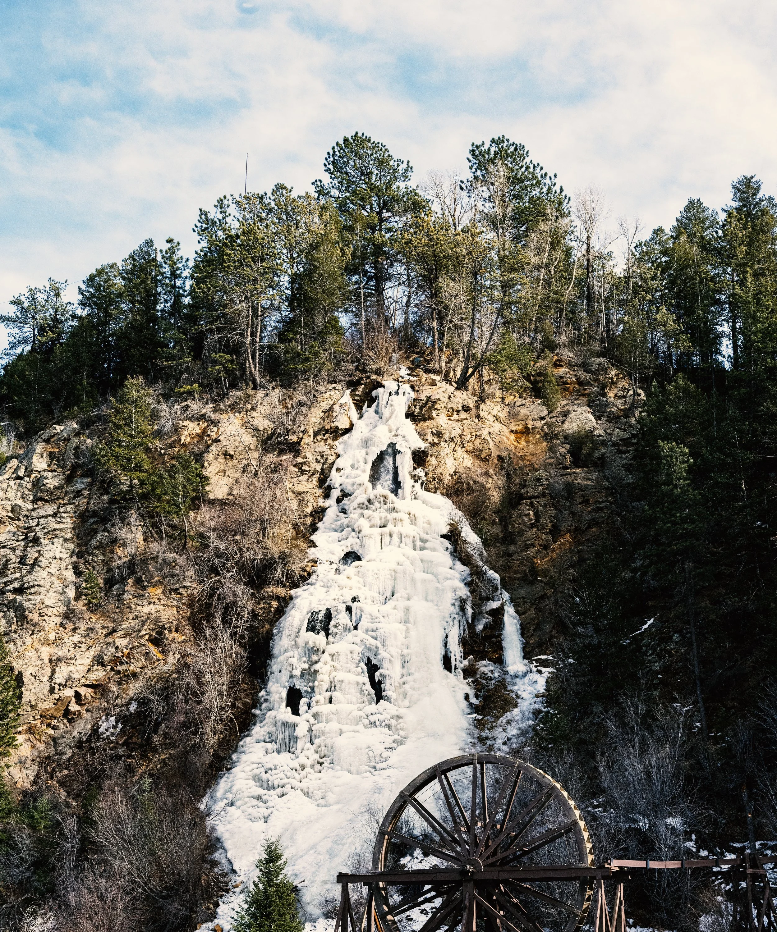 A waterfall frozen in ice flowing down a rocky hillside surrounded by trees, with part of an old wooden water wheel in the foreground and a cloudy sky in the background.