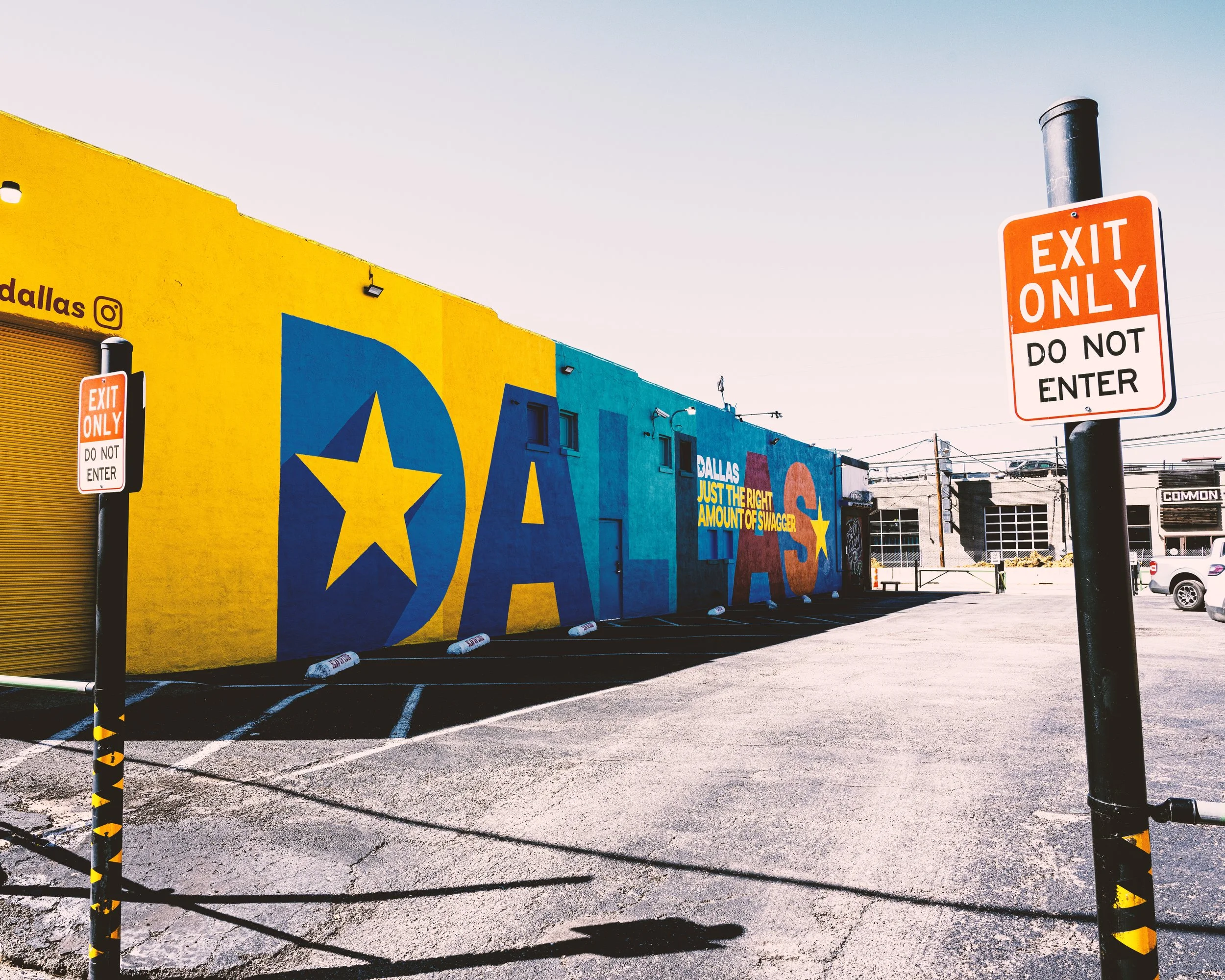 A colorful mural on a yellow and blue building with a blue star and yellow star. In the foreground, there are two orange and white signs that say "Exit Only" and "Do Not Enter". The parking lot is mostly empty with a few cars and buildings in the bac