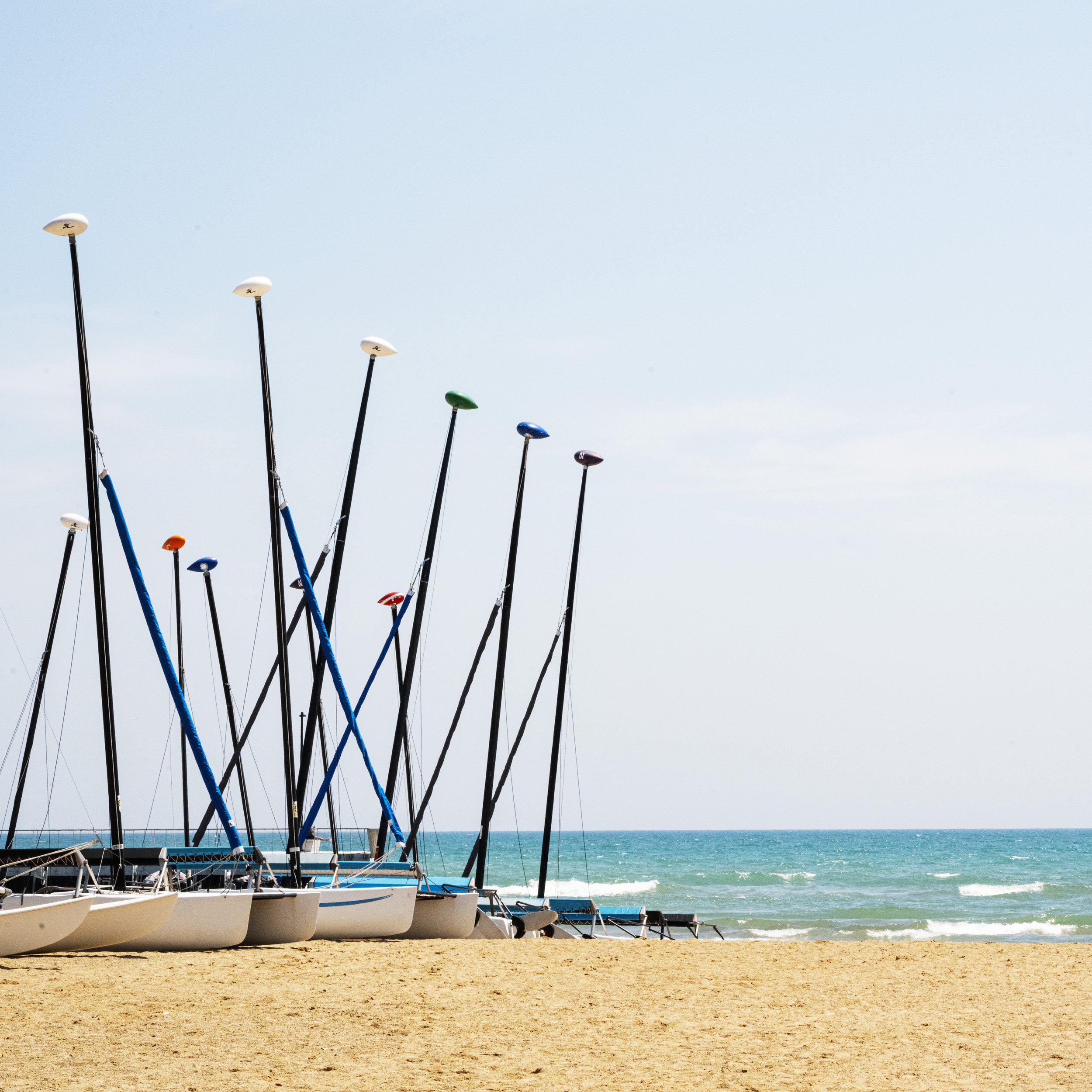 A row of sailboats with colorful masts and sails, resting on a sandy beach near the ocean, with waves and a blue sky in the background.