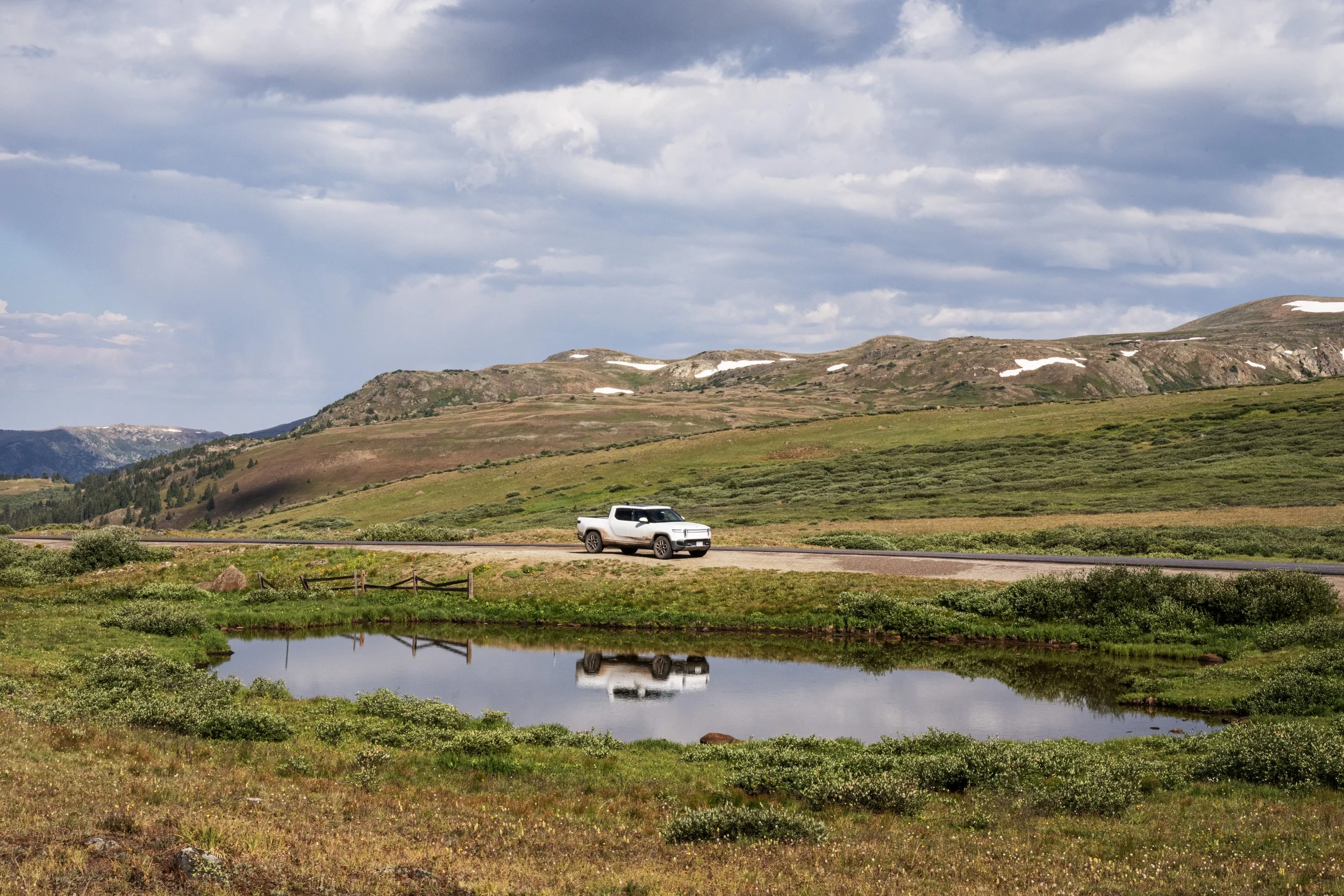 A white pickup truck traveling on a curved road in a rural landscape with green grass, small pond, and rolling hills with patches of snow under a cloudy sky.