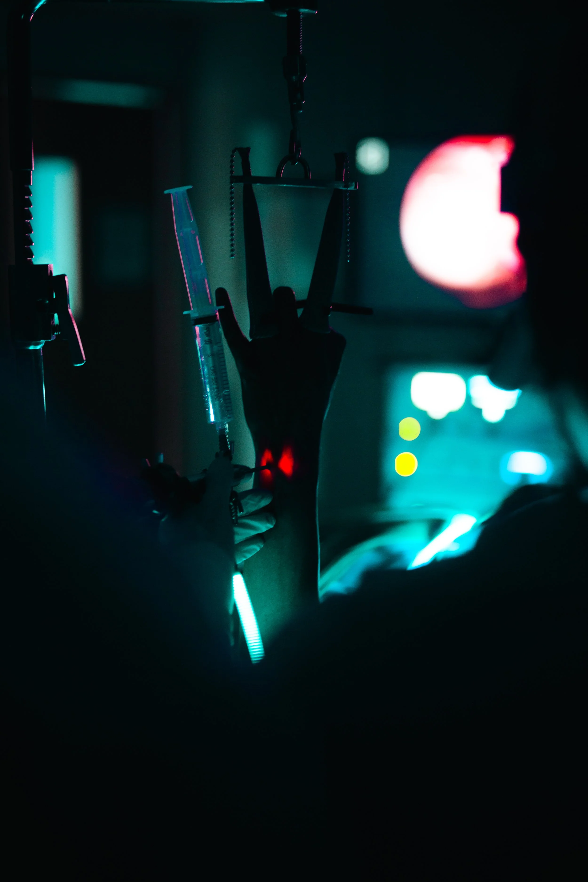 Mano siendo atendida con una inyección en un entorno médico oscuro, con luces de colores y equipo médico visible.