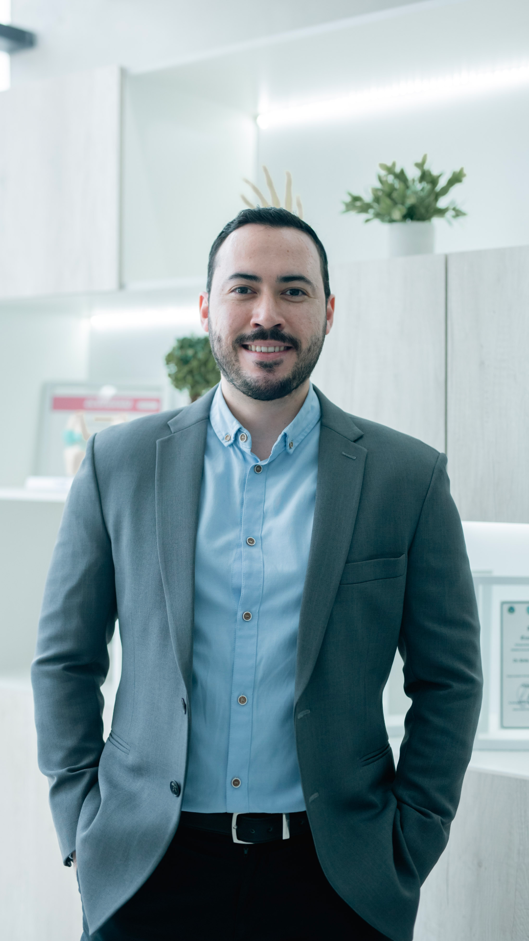 Hombre sonriendo, vestido con saco gris y camisa azul claro, en un entorno profesional con plantas decorativas.
