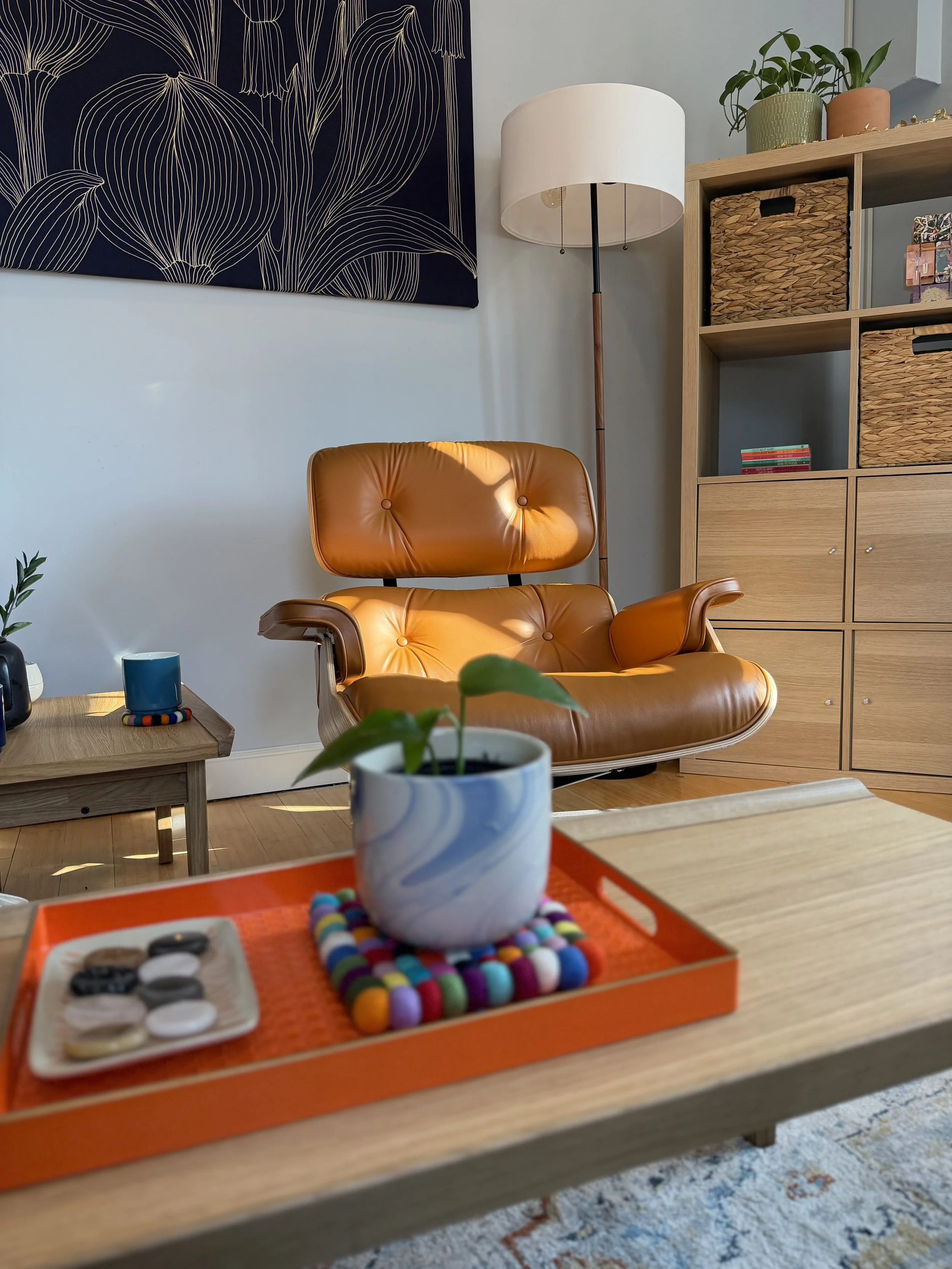 Living room with a tan leather lounge chair, a wooden shelf with baskets and plants, a floor lamp, a multi-colored tray with a ceramic pot and small items on a wooden table, and a large black and white botanical wall art.