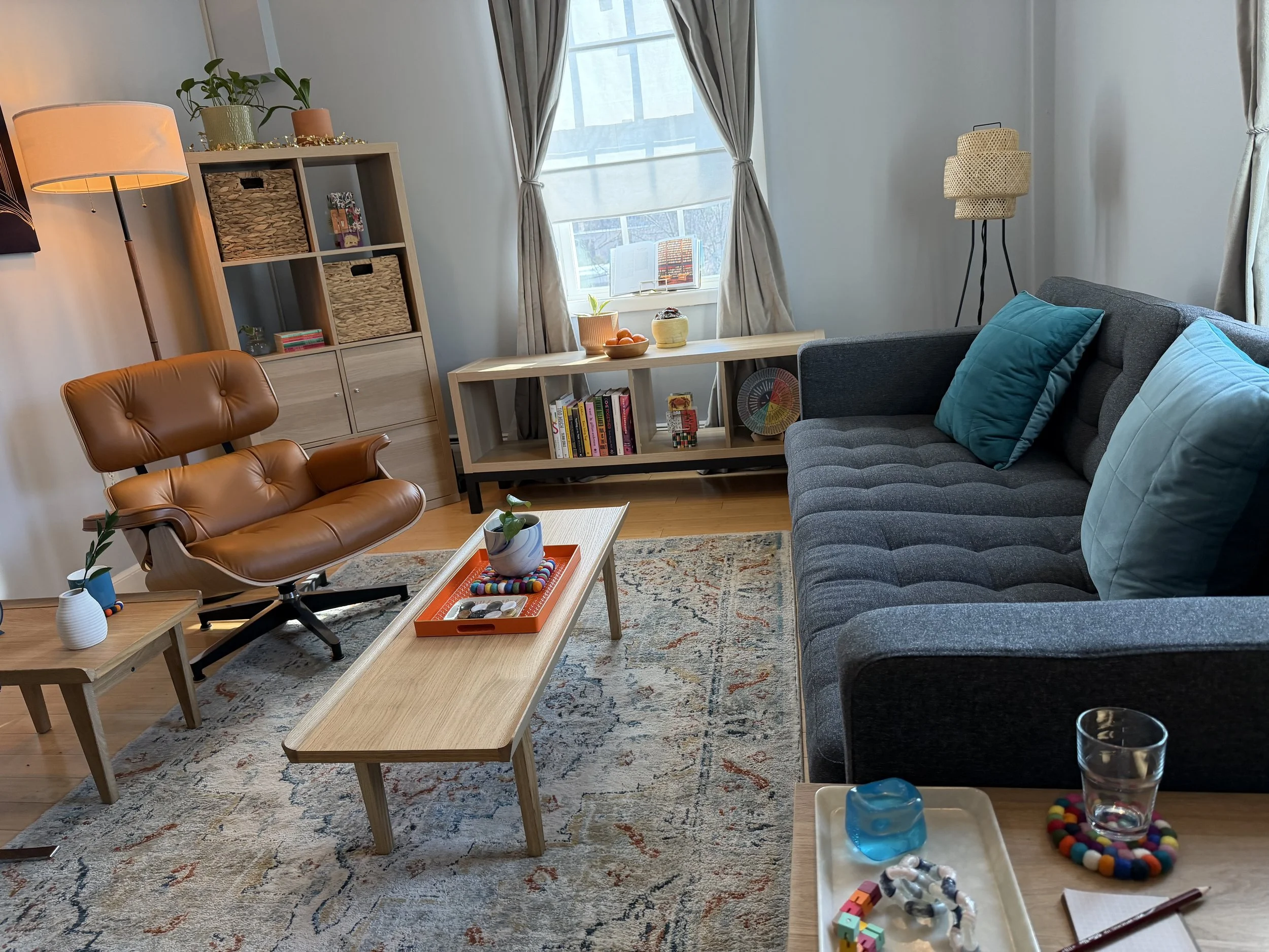 Living room with blue sofa, teal and light blue pillows, brown leather chair, wooden coffee table, patterned rug, bookshelf, side table with glass, and window with curtains.