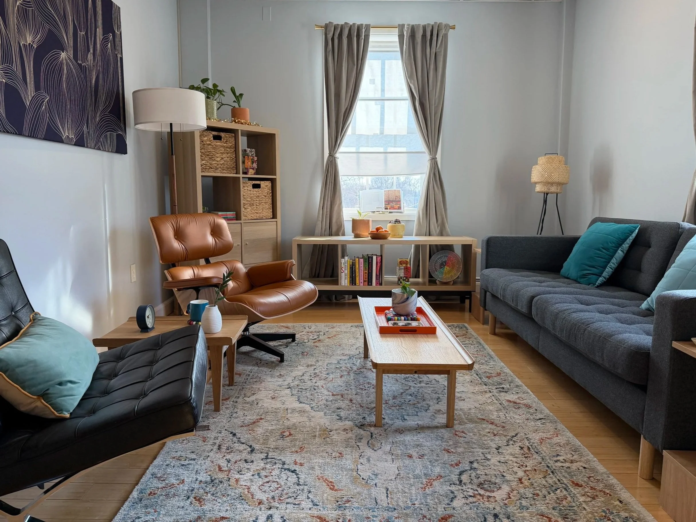 Living room with black leather and gray fabric sofas, a wooden coffee table with decorative items, a patterned rug, a window with beige curtains, and a bookshelf with baskets and books.