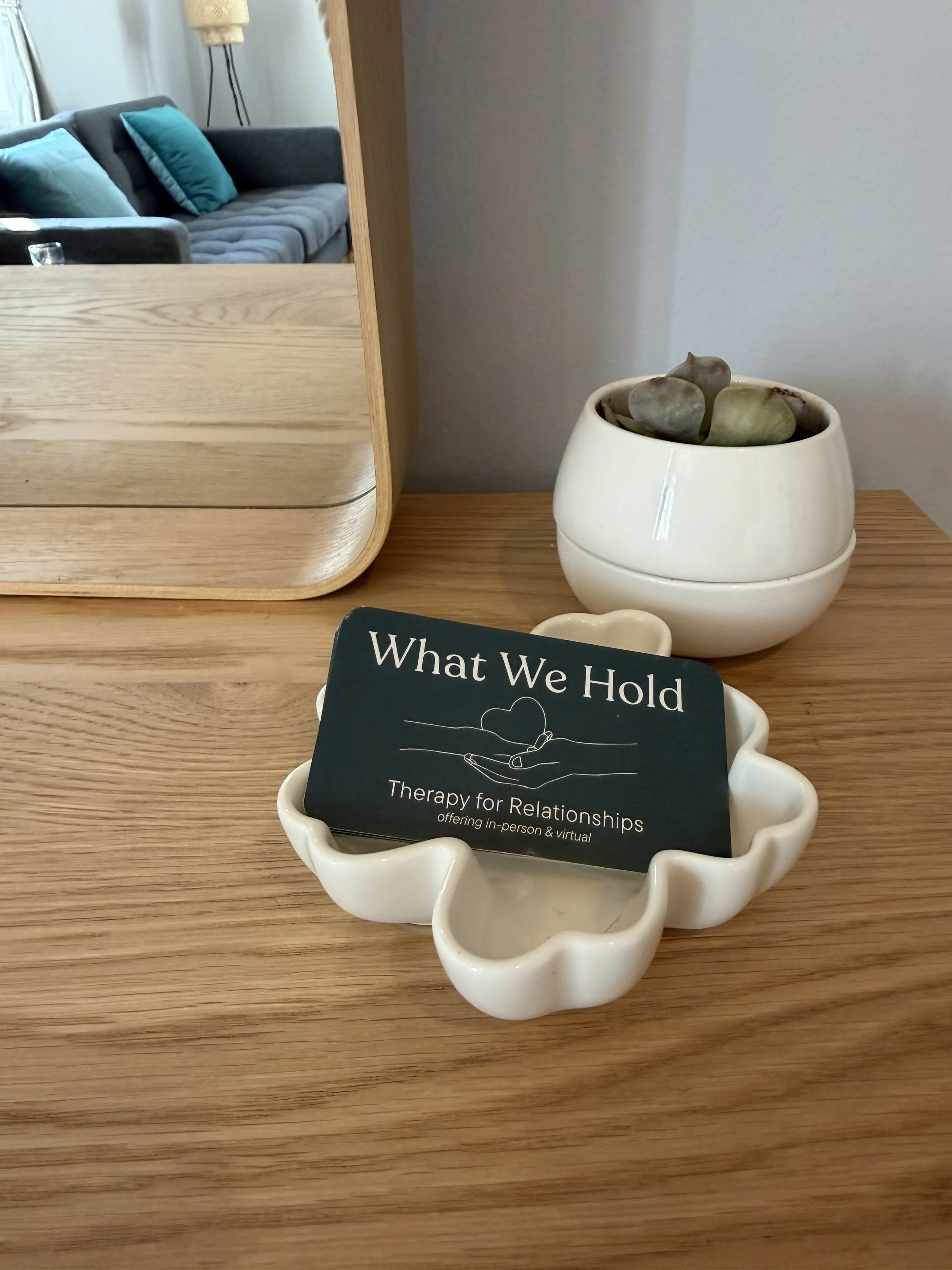 A wooden table with a decorative white shell-shaped dish holding a business card that reads 'What We Hold, Therapy for Relationships, offering in-person & virtual.' Next to the dish is a round white ceramic planter with succulents. A mirror on the le