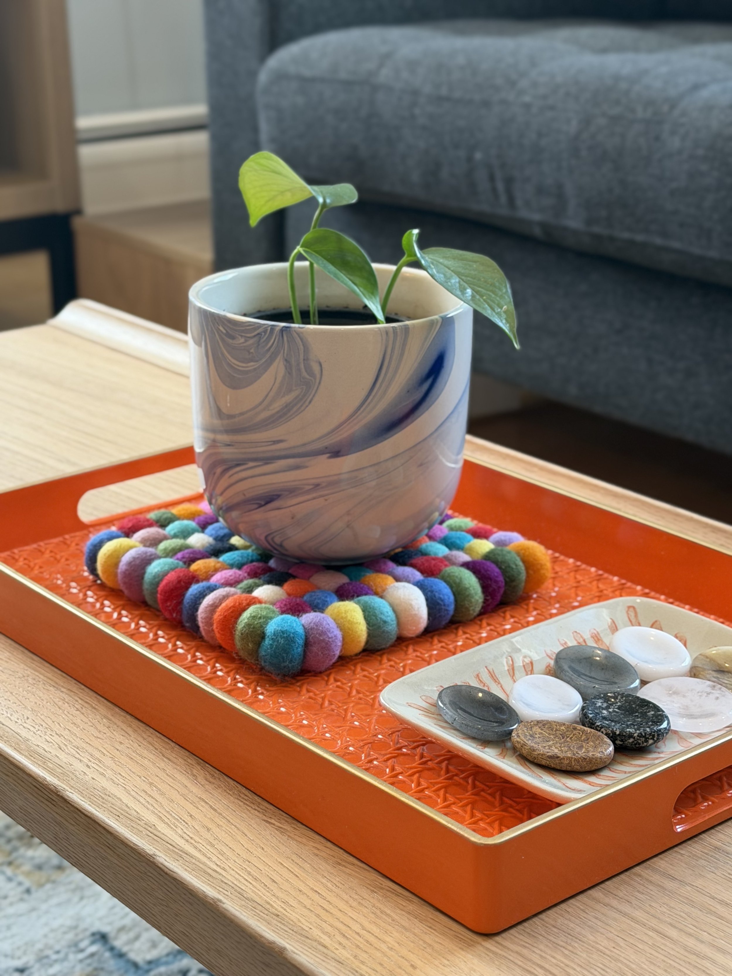 A decorative indoor setup with a small potted plant on a colorful felt mat, placed on an orange tray with a white rectangular dish containing assorted smooth, polished stones.