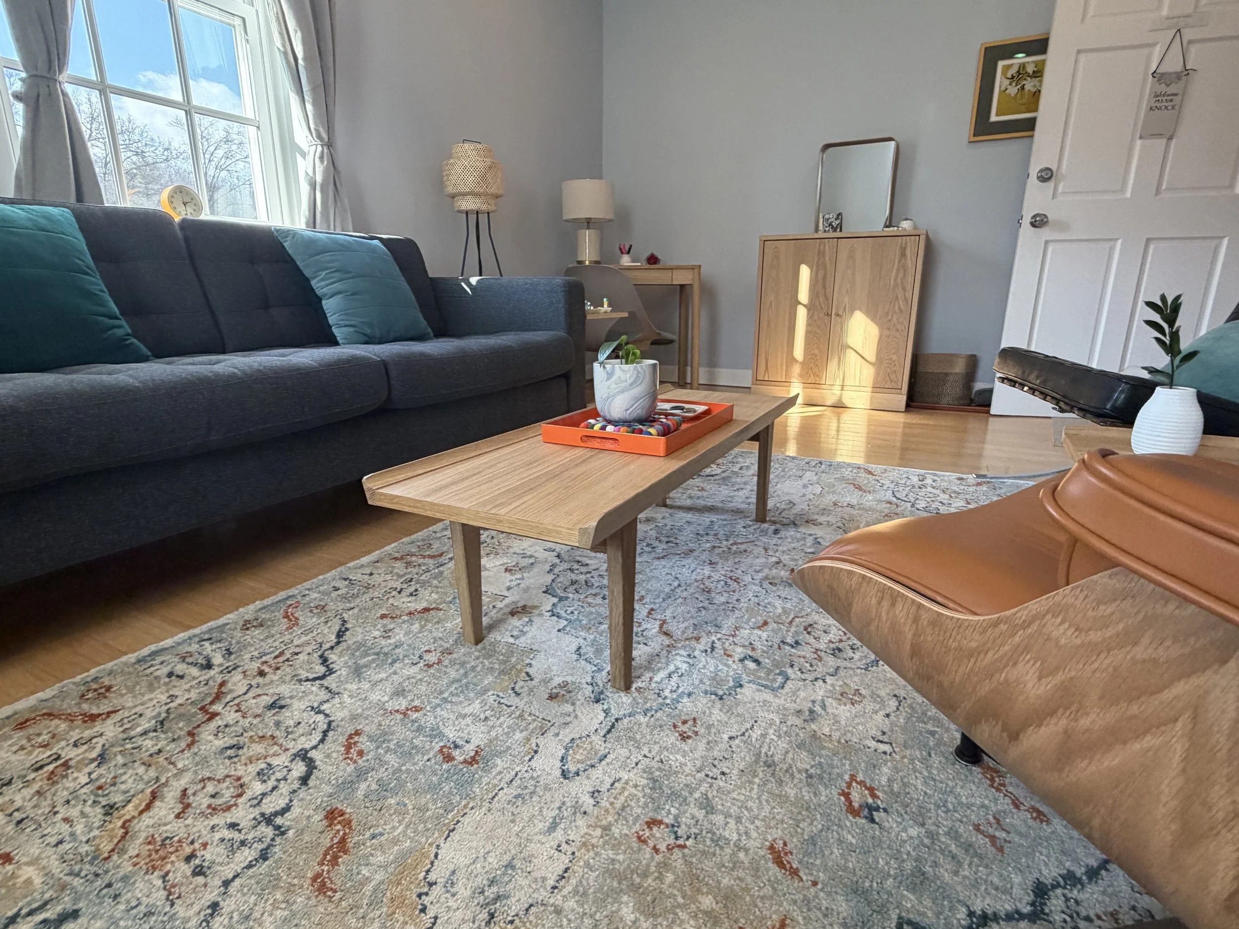 Living room with gray couch, wooden coffee table, patterned rug, armchair, and decor including pillows, vases, and artwork, with natural light coming through a window.