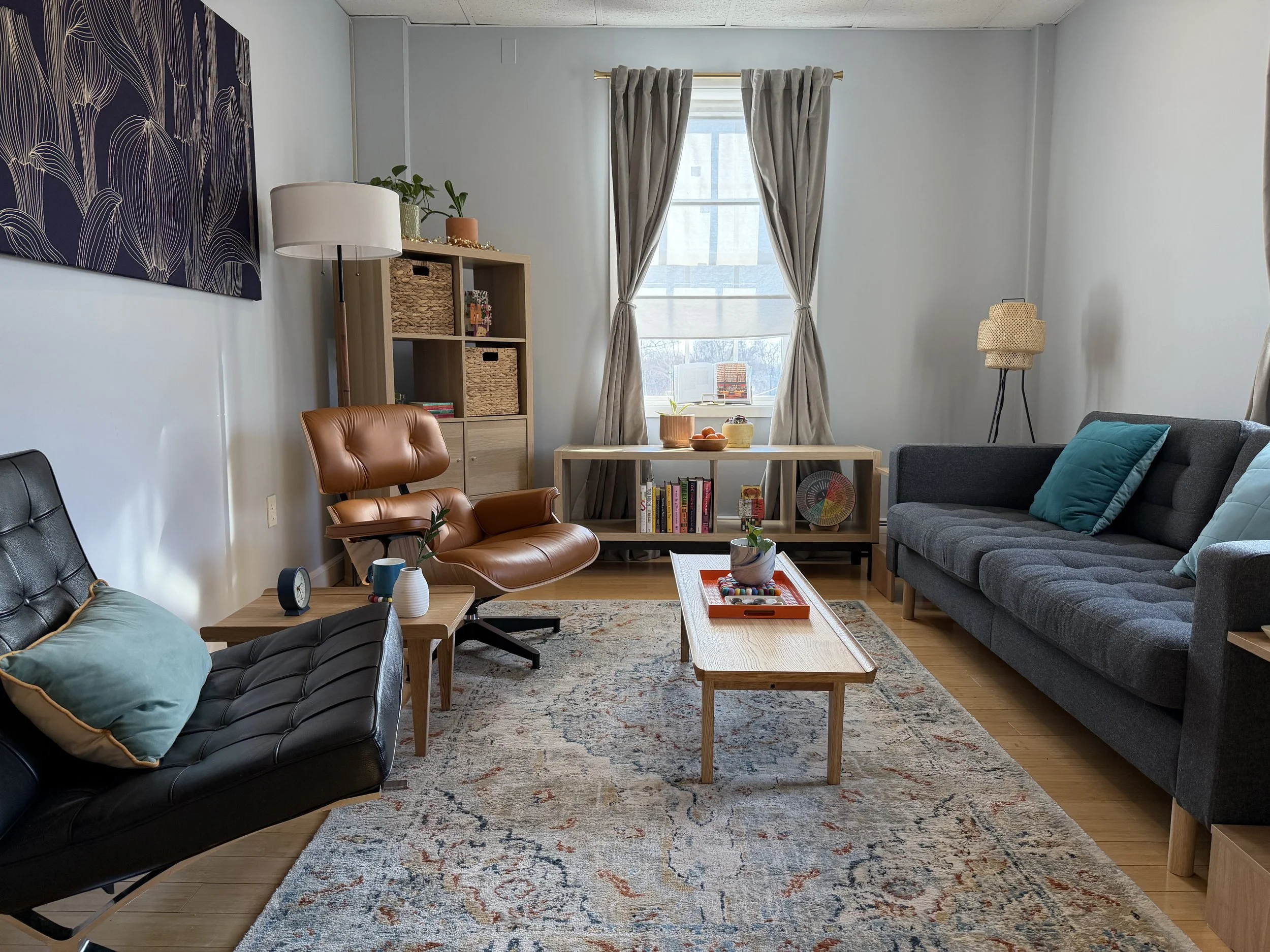 Living room with black leather and gray fabric sofas, a wooden coffee table with decorative items, a patterned rug, a window with beige curtains, and a bookshelf with baskets and books.