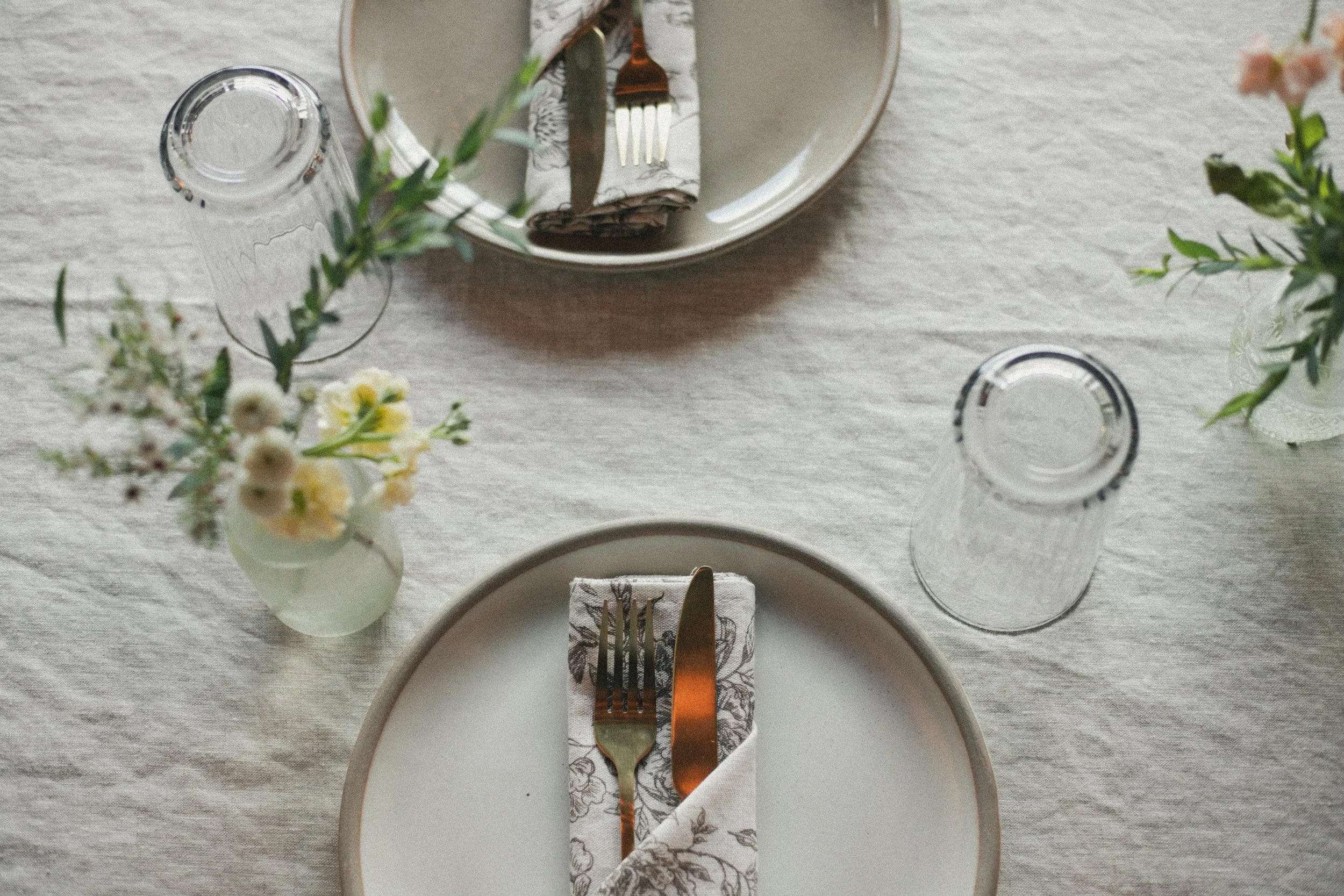 A table setting with a white plate, cutlery wrapped in a napkin, two clear empty glasses, and small floral arrangements in vases.