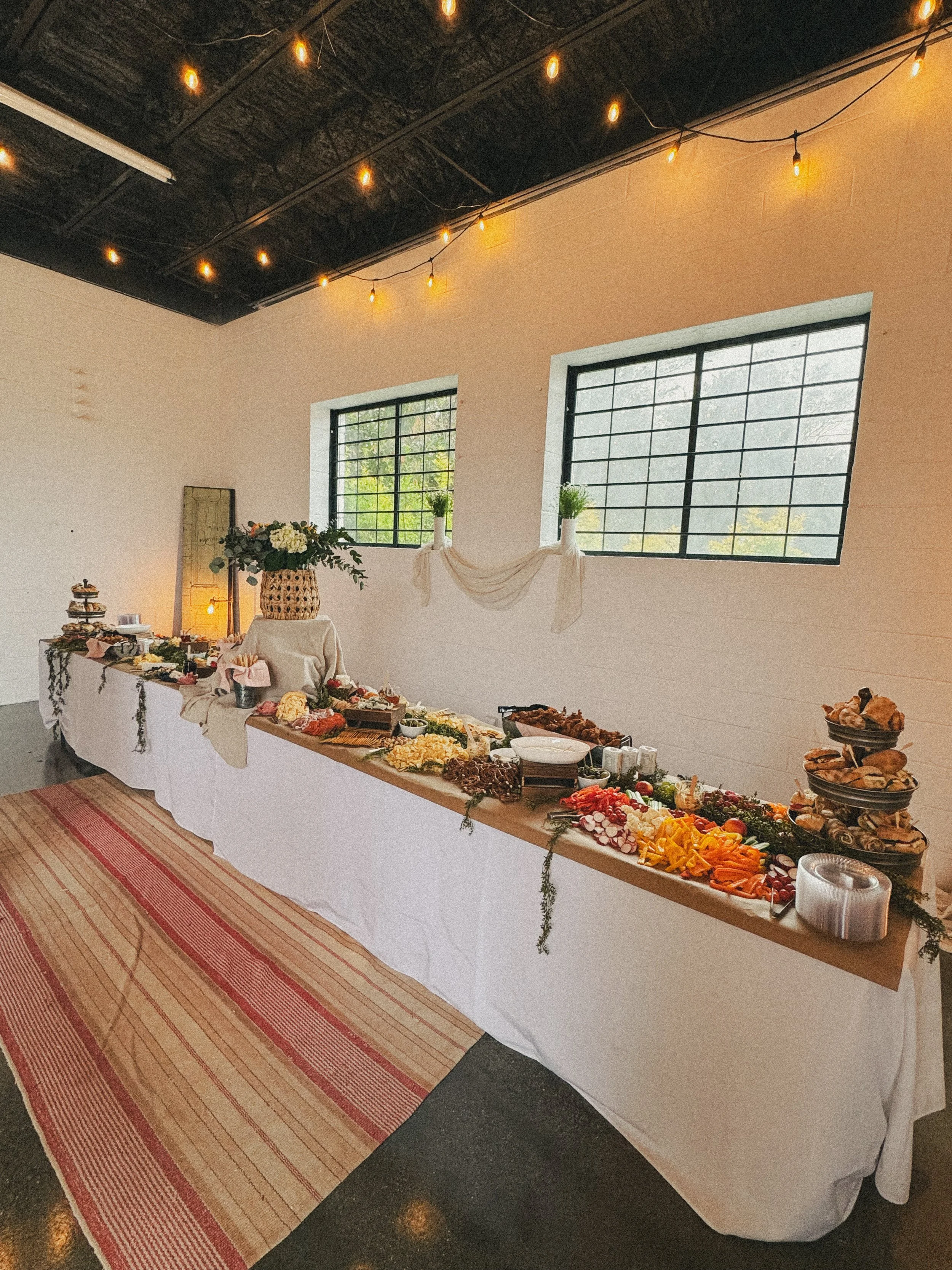 A long buffet table covered with a white tablecloth, adorned with flowers and decorated greenery, set in a room with large windows and string lights on the ceiling. The table displays various foods, including vegetables, cheeses, and baked goods.