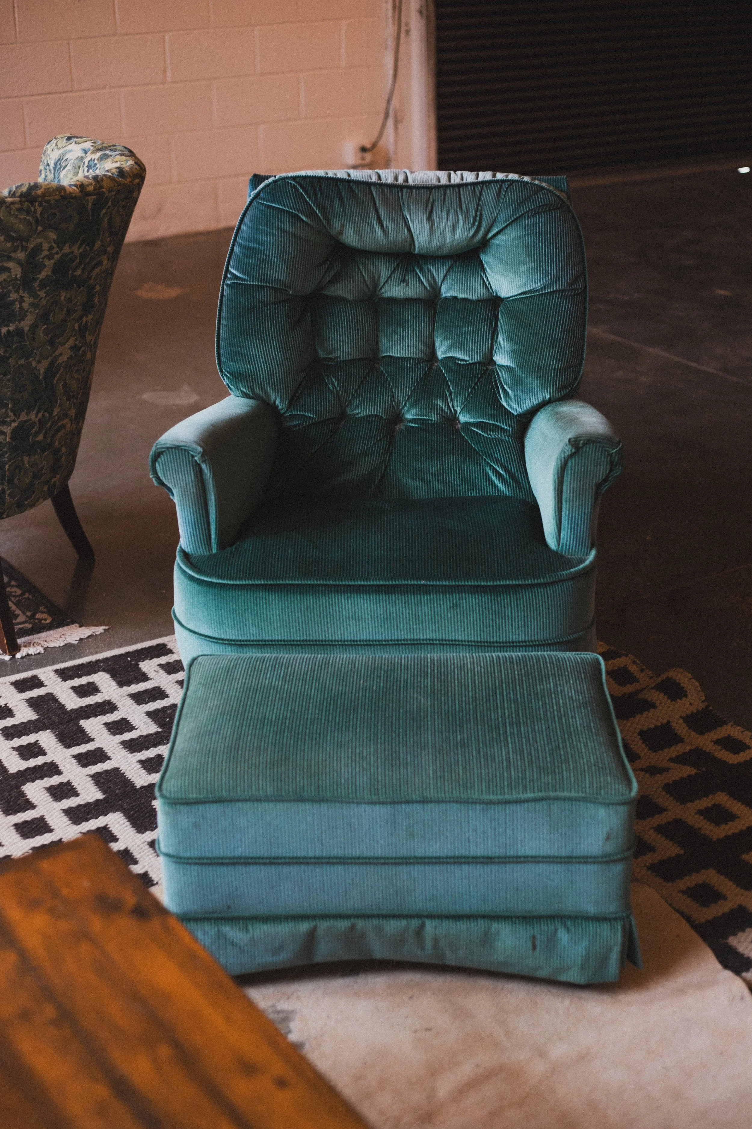 A teal upholstered armchair with a matching ottoman in a room with a wooden floor and a patterned area rug.