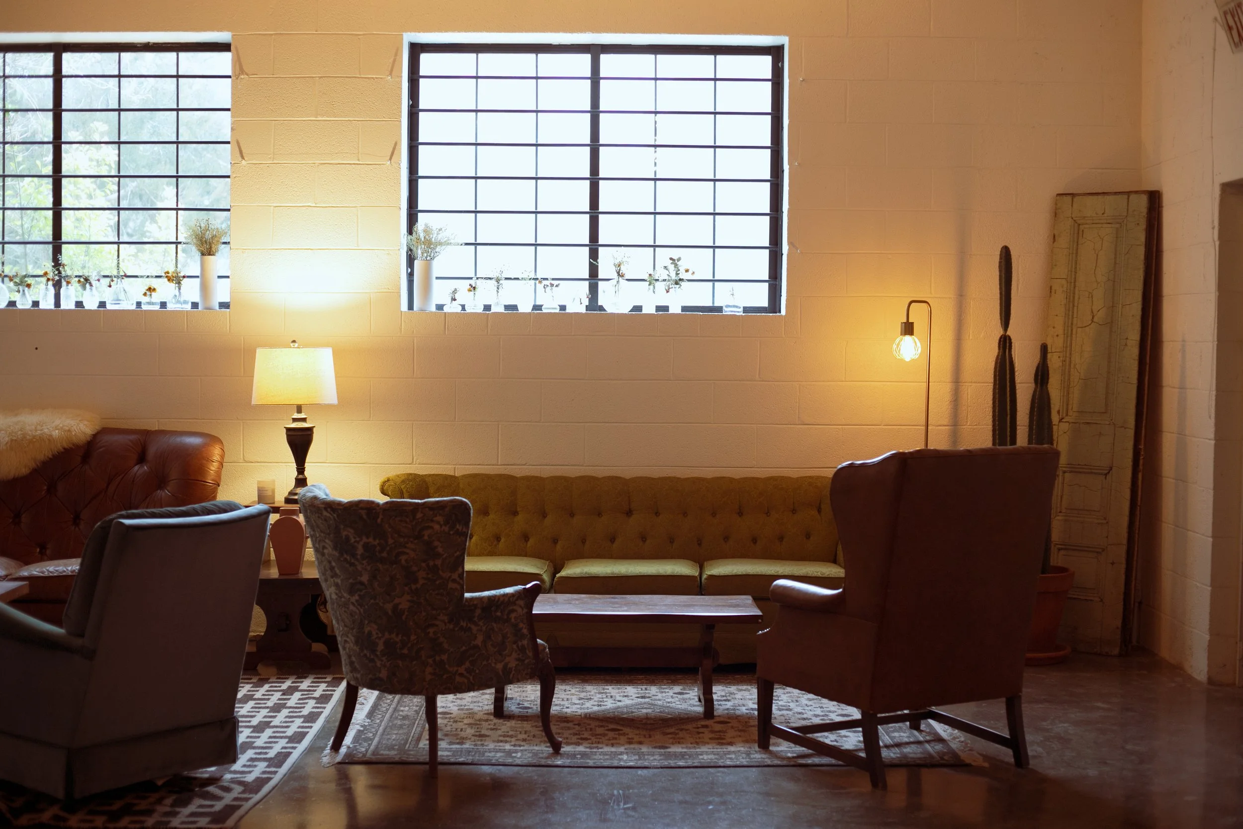 Interior of a cozy living room with two large windows, a yellow tufted sofa, a patterned armchair, and various lamps providing warm lighting.