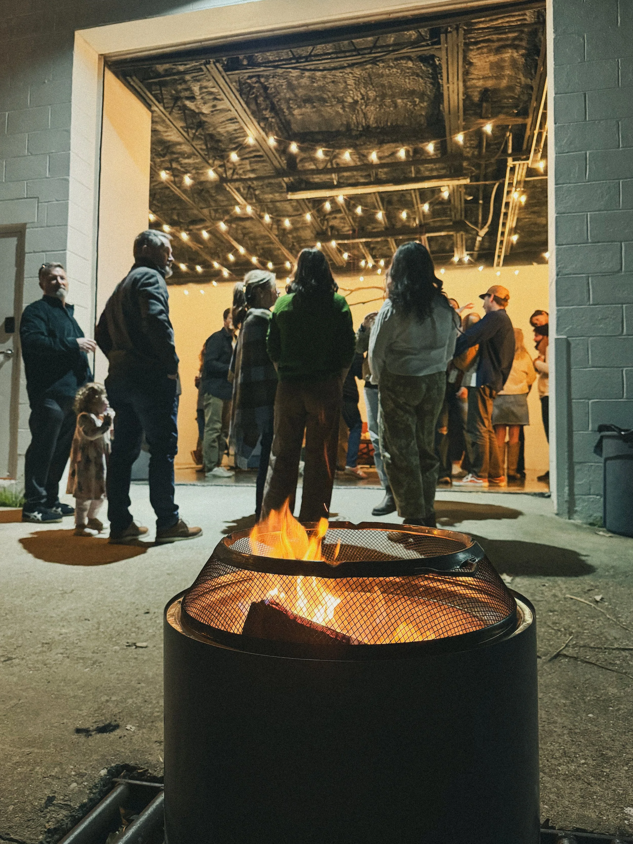 A group of people gathered inside a building with a ceiling decorated with string lights, standing near a fire pit outdoors at night, viewed from behind.