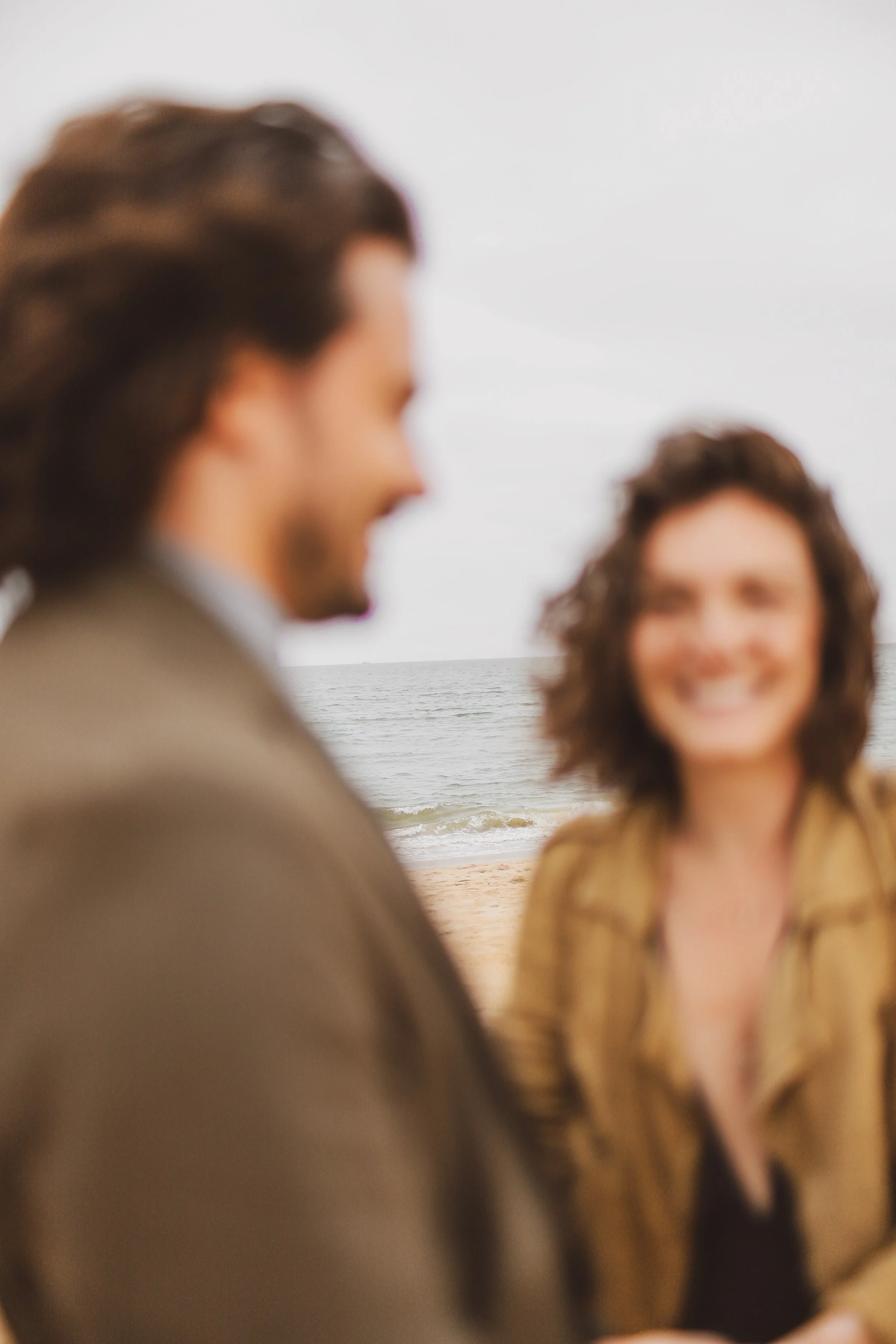 Two people are standing on a beach, blurred in the foreground, with the ocean in the background. The woman has curly hair and is smiling, while the man has longer hair and a beard, with his face partially turned away.