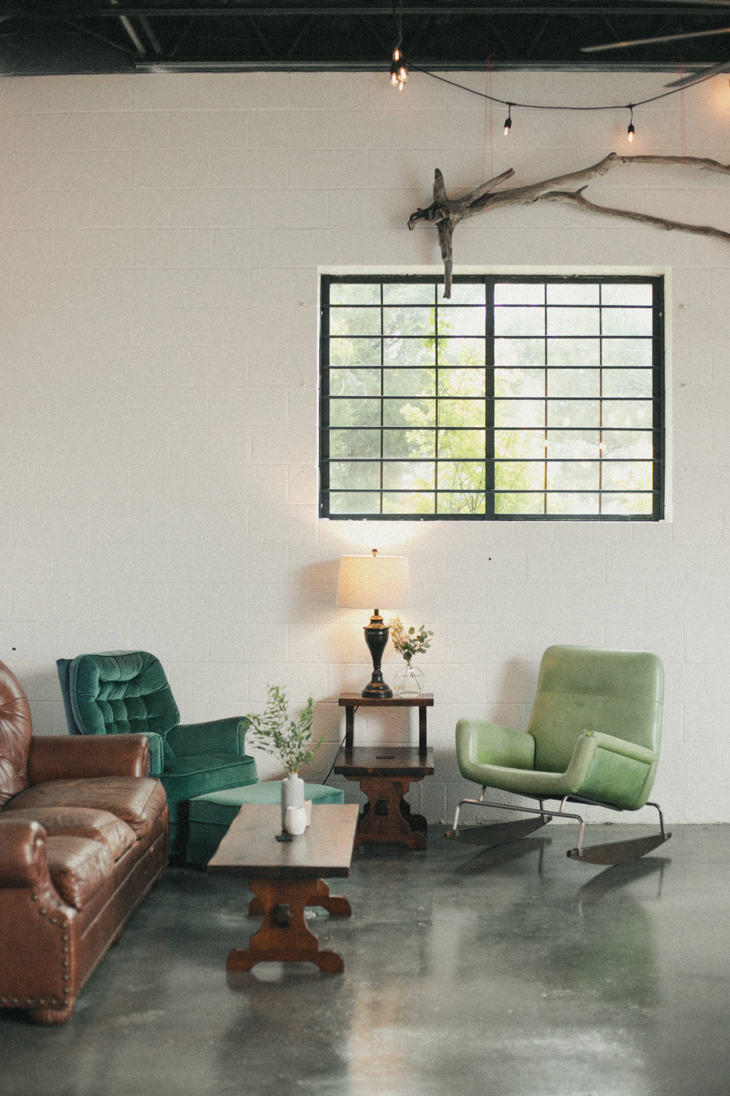 Interior of a minimalist living room with vintage and modern furniture, including green and brown leather chairs, a wooden coffee table, a side table with a lamp, and a large window with metal bars, decorated with hanging string lights and a driftwoo