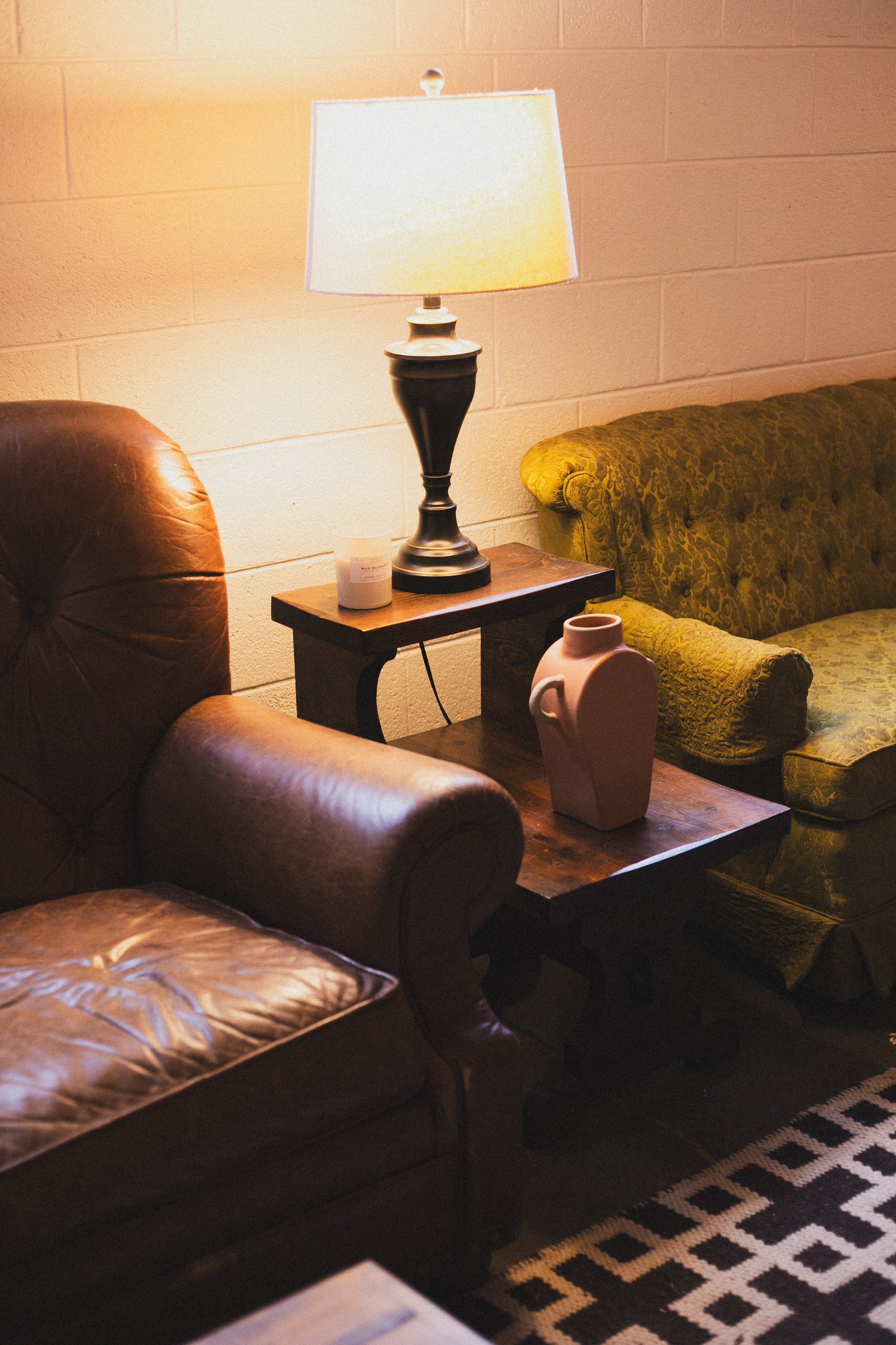 A cozy living room corner with a leather armchair and a patterned rug. A wooden side table holds a lamp with a black base and white shade, a small candle, and a decorative pink vase. A green patterned couch is partially visible in the background with
