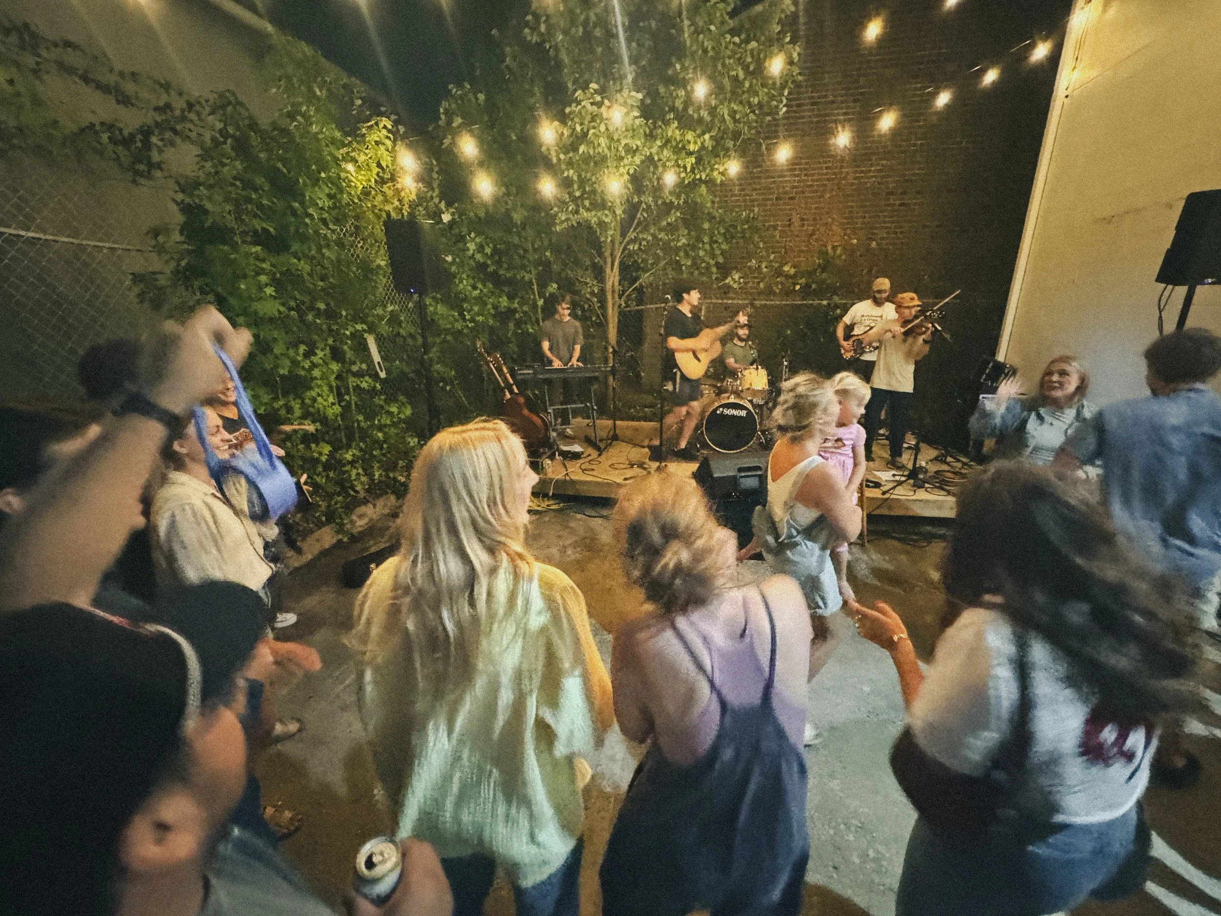 People dancing and enjoying live music at an outdoor event during the evening, with a band playing on stage under string lights and trees.