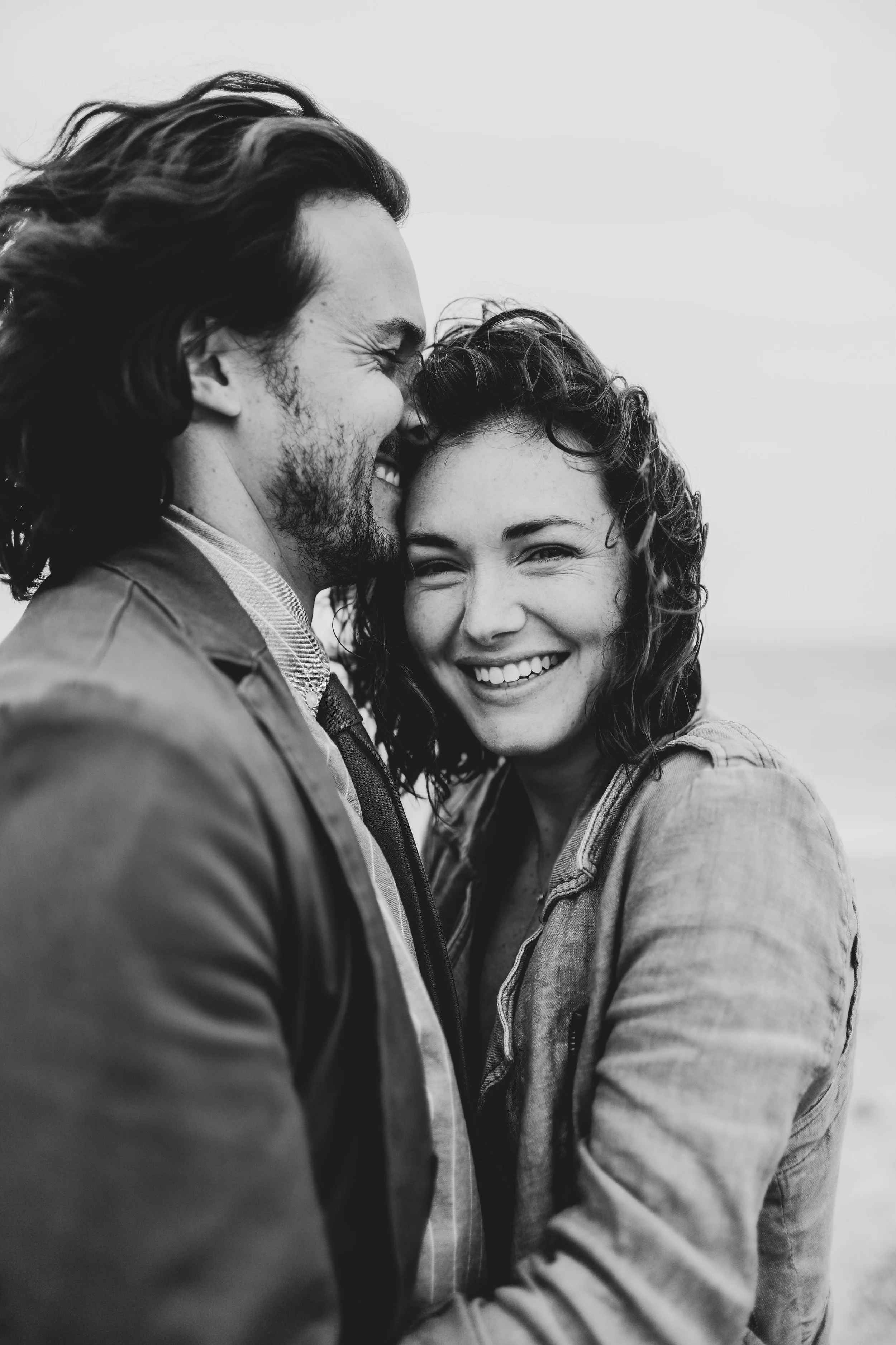 A black and white photo of a young man and woman warmly smiling, with their foreheads touching and eyes closed, showing affection and joy.