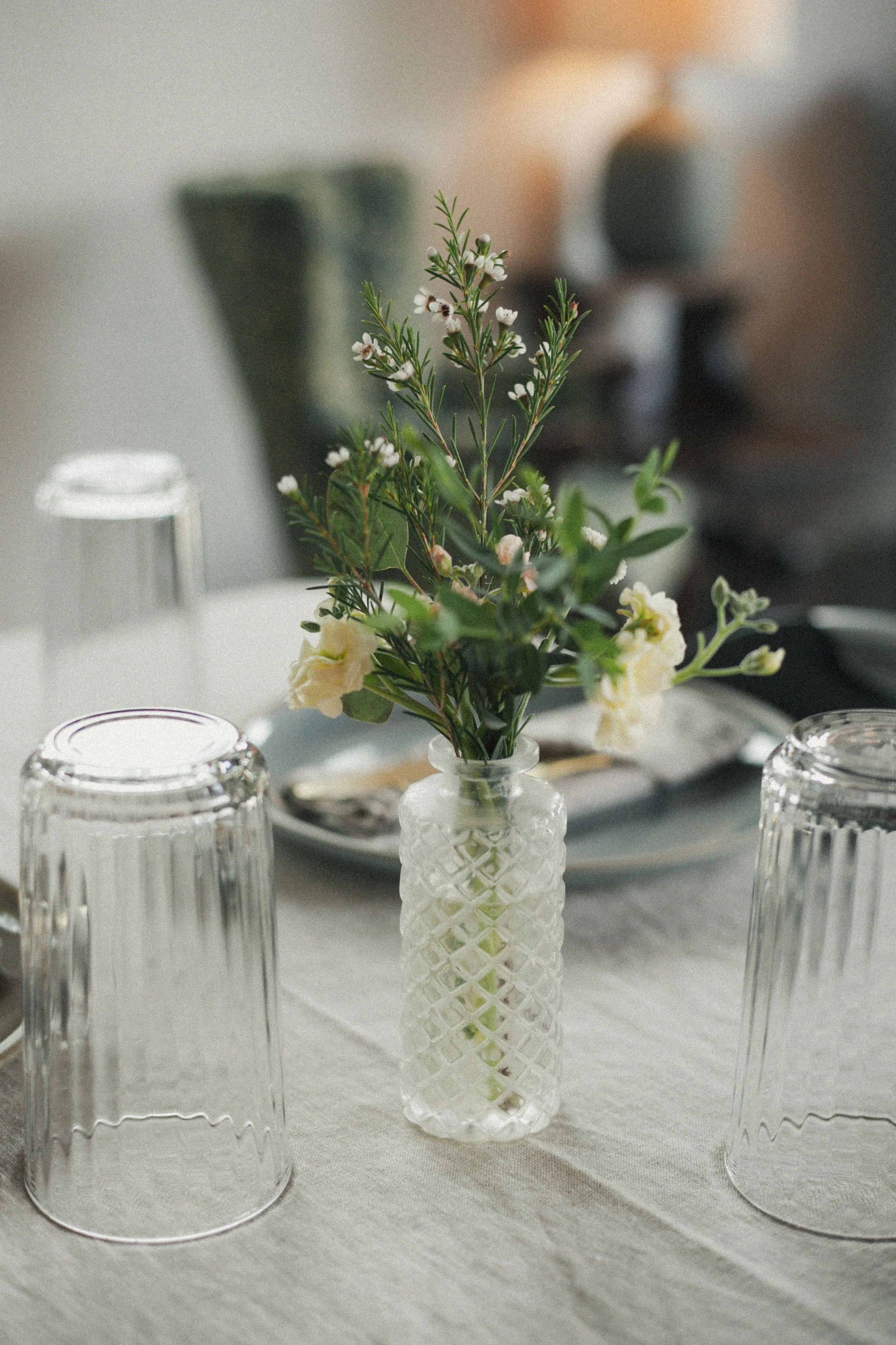 A table setting with a white lattice-patterned glass vase holding green foliage and white flowers, surrounded by upside-down clear glass tumblers, on a white tablecloth in a softly lit room.