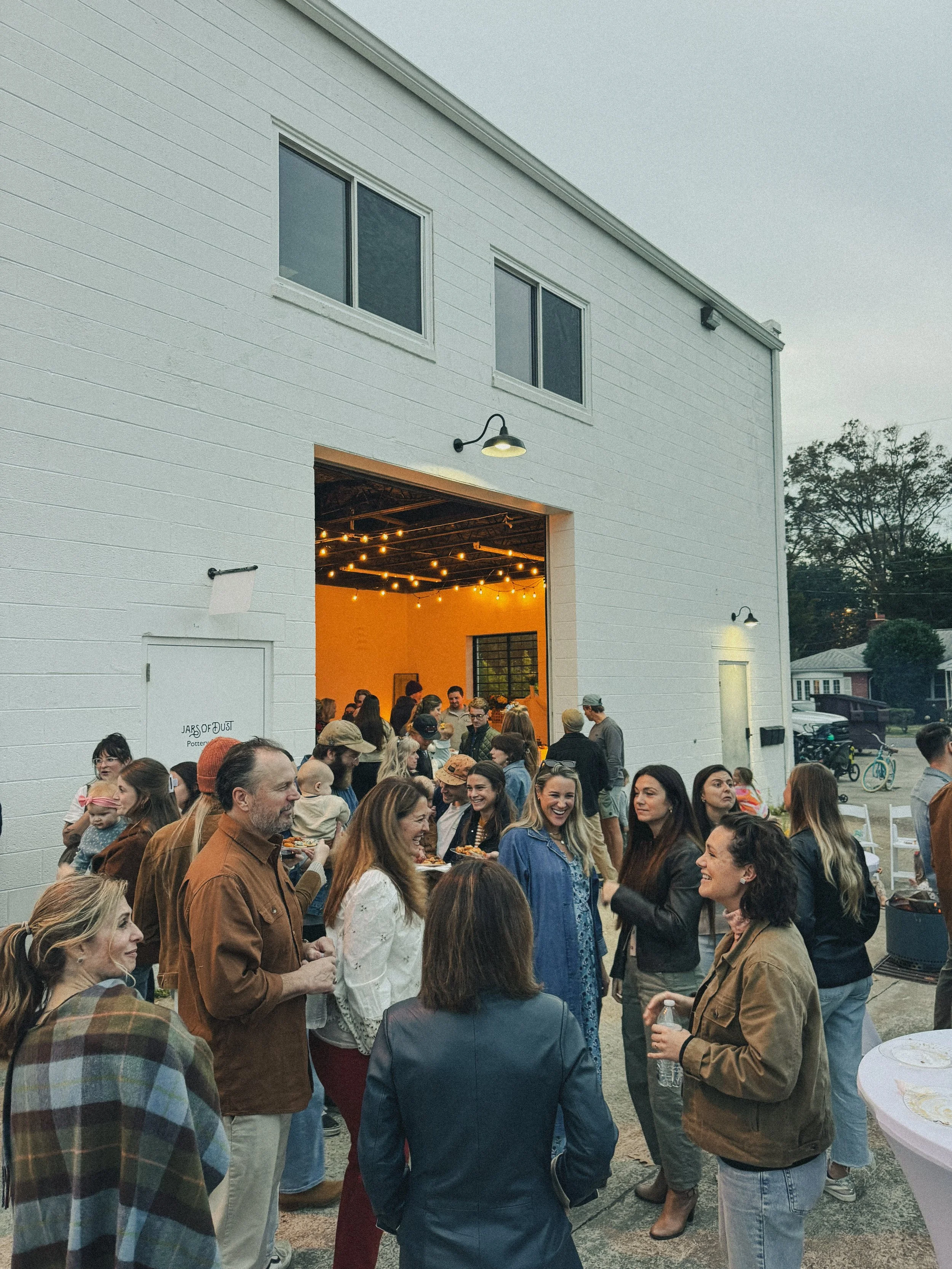 A gathering of people outside a white building with large open garage door, hanging string lights, and windows, during evening or night time.
