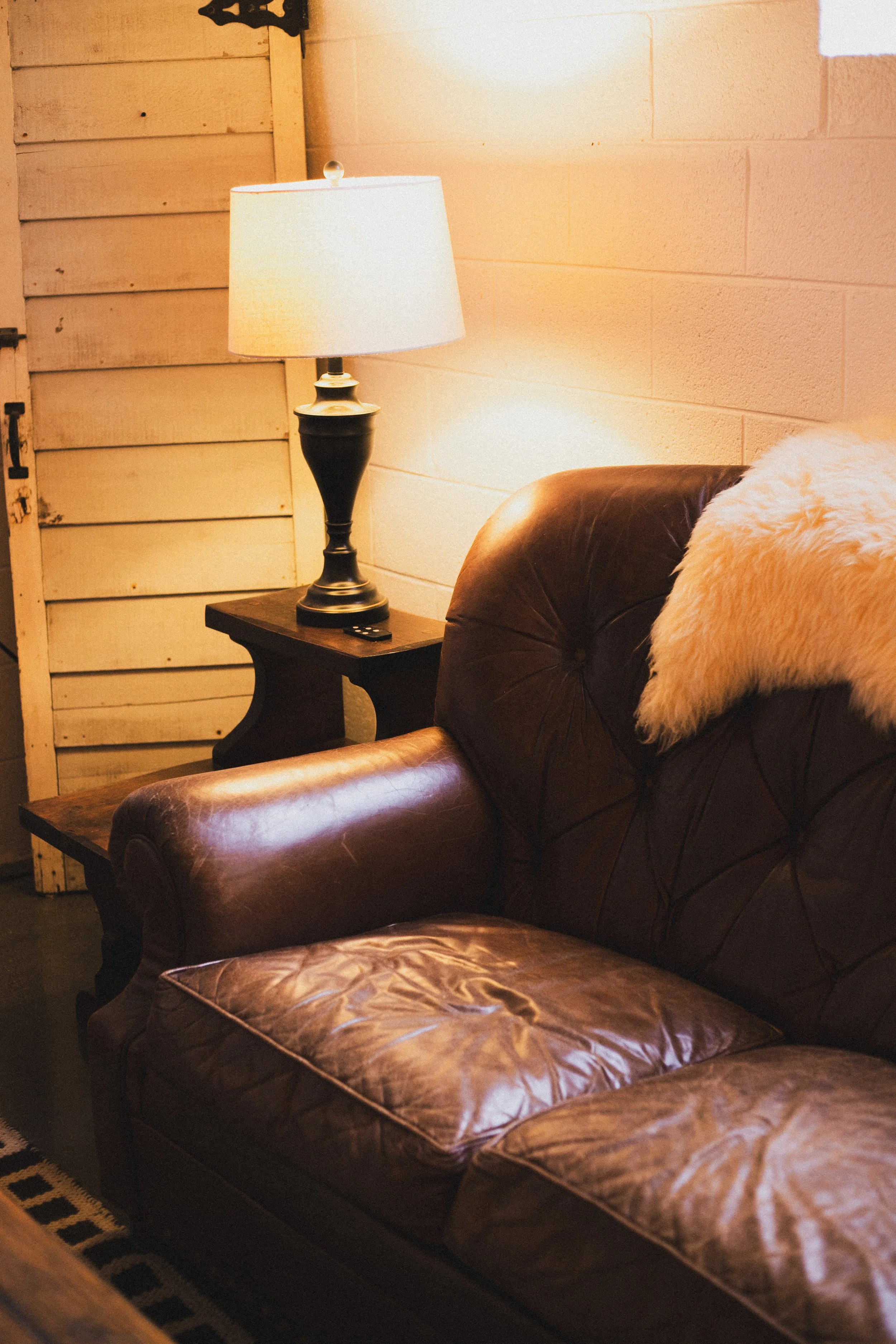 A cozy living room corner with a leather sofa, plush cream pillow, wooden side table with a lamp, and a textured wooden wall background.