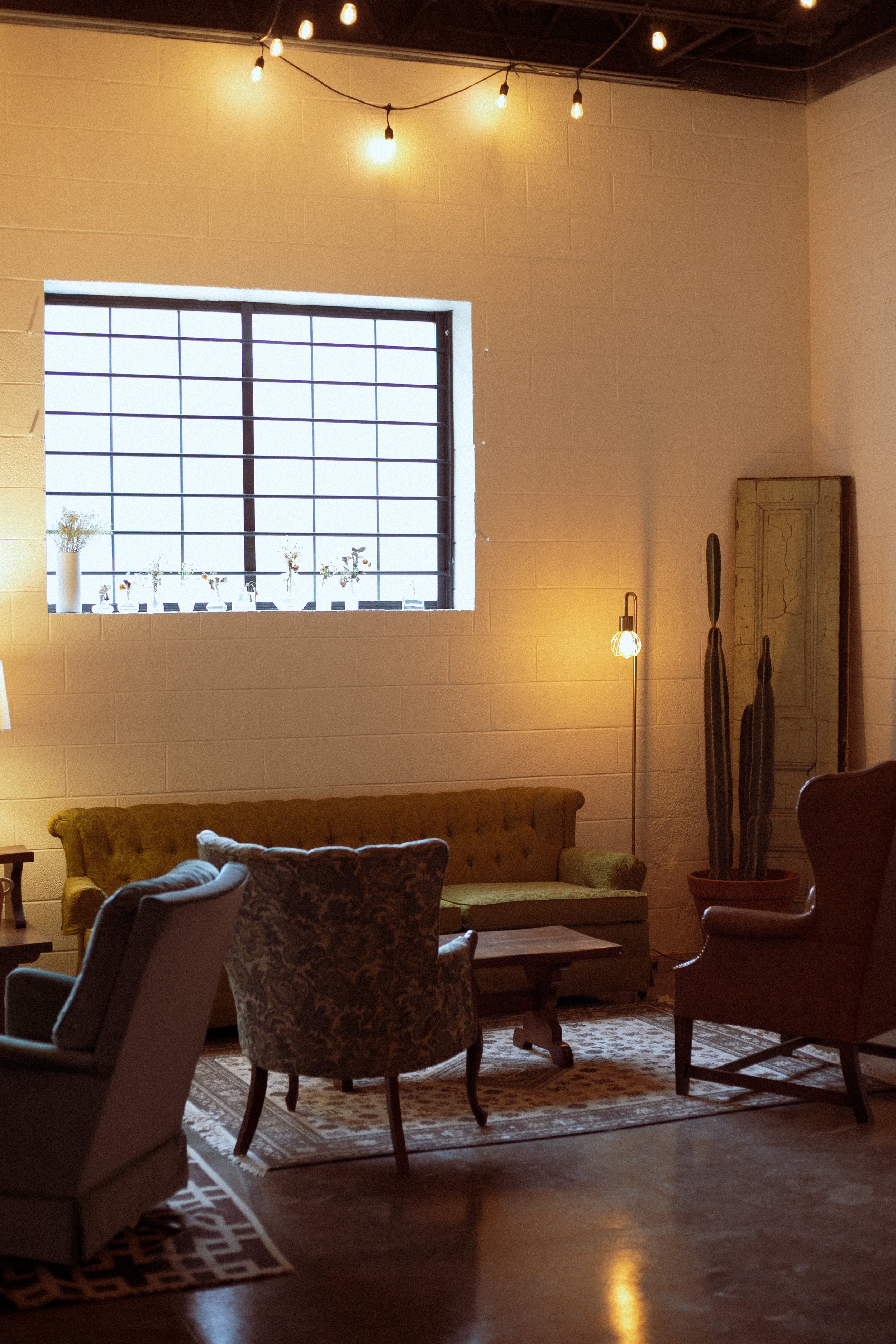 A cozy living room with a large window, string lights hanging from the ceiling, and various vintage and modern chairs and a green tufted sofa arranged around a wooden coffee table. There's a tall potted cactus, a floor lamp with a visible bulb, and a