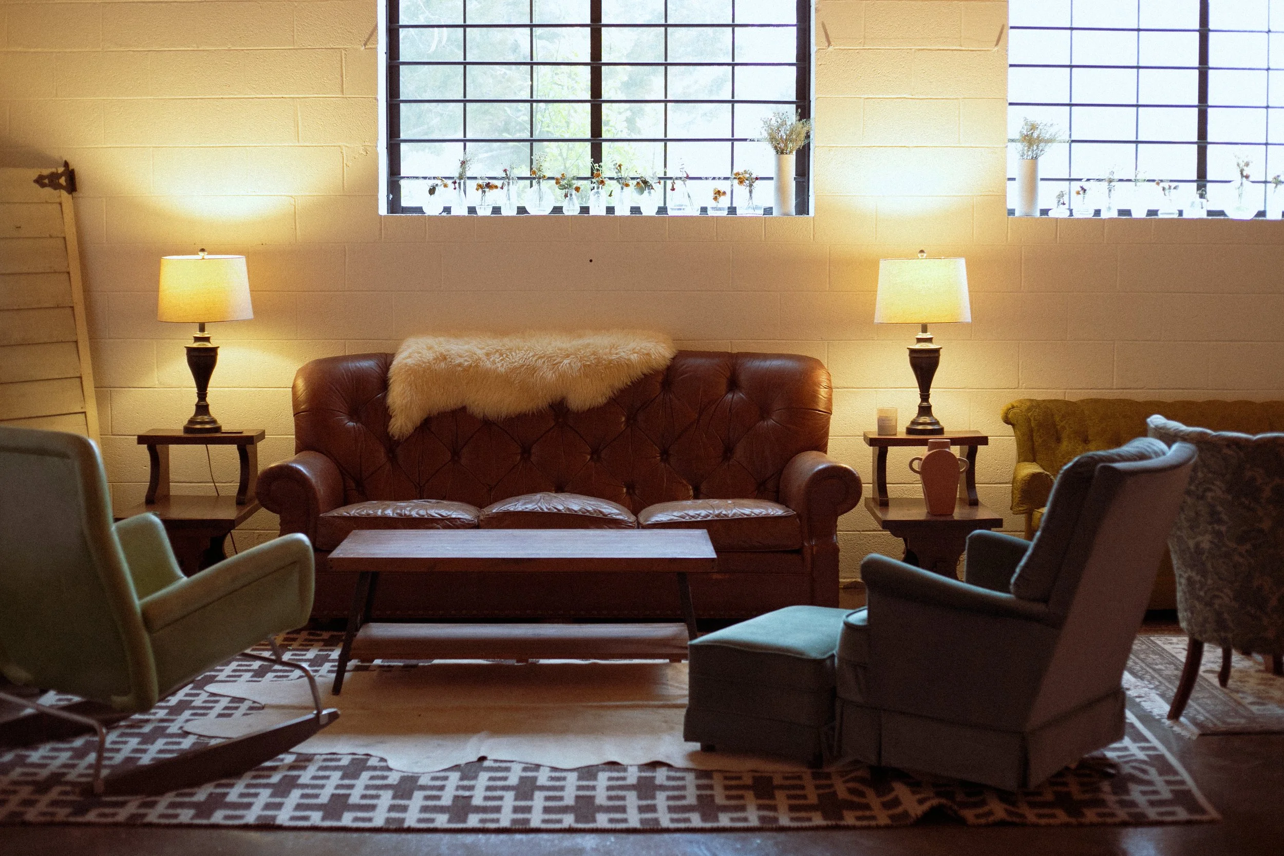 A cozy living room with a brick wall, two windows with black frames, and various vases with flowers on the windowsill. There are two lamps on side tables, a leather couch, an armchair, a wooden coffee table, and a patterned rug.