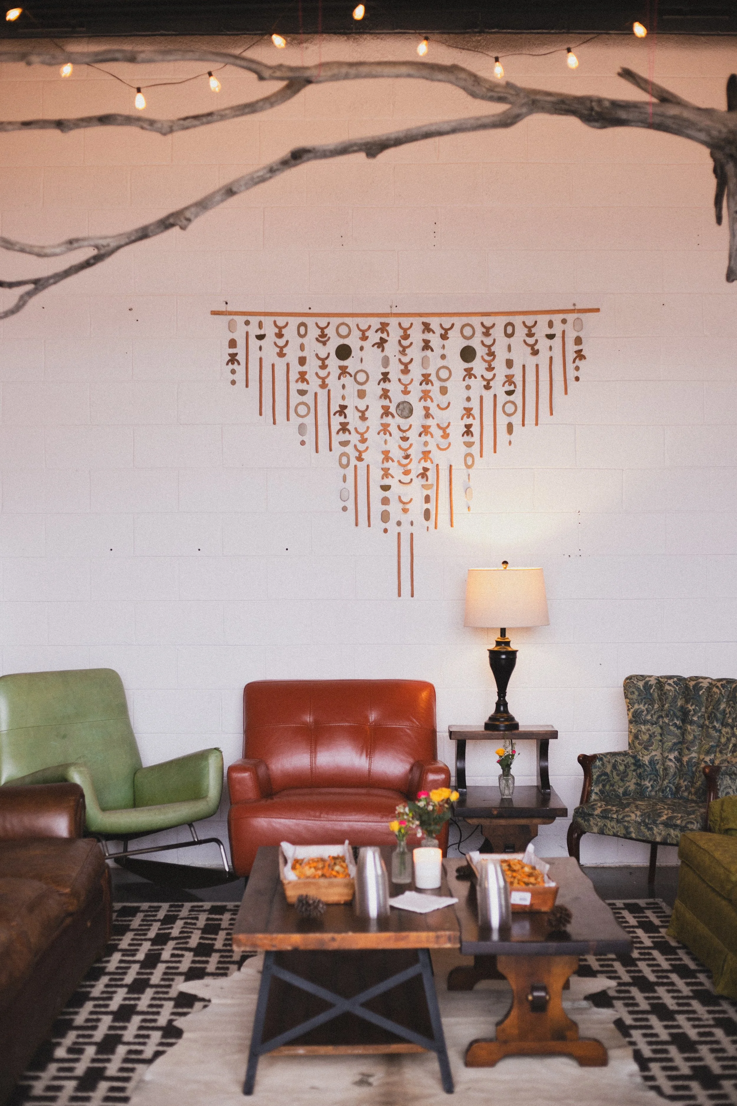 Cozy living room with colorful armchairs, a wooden coffee table, and a black-and-white geometric patterned rug. There's a lamp with a cream shade on a side table, a decorative wall hanging, and string lights hanging from above. A small flower arrange
