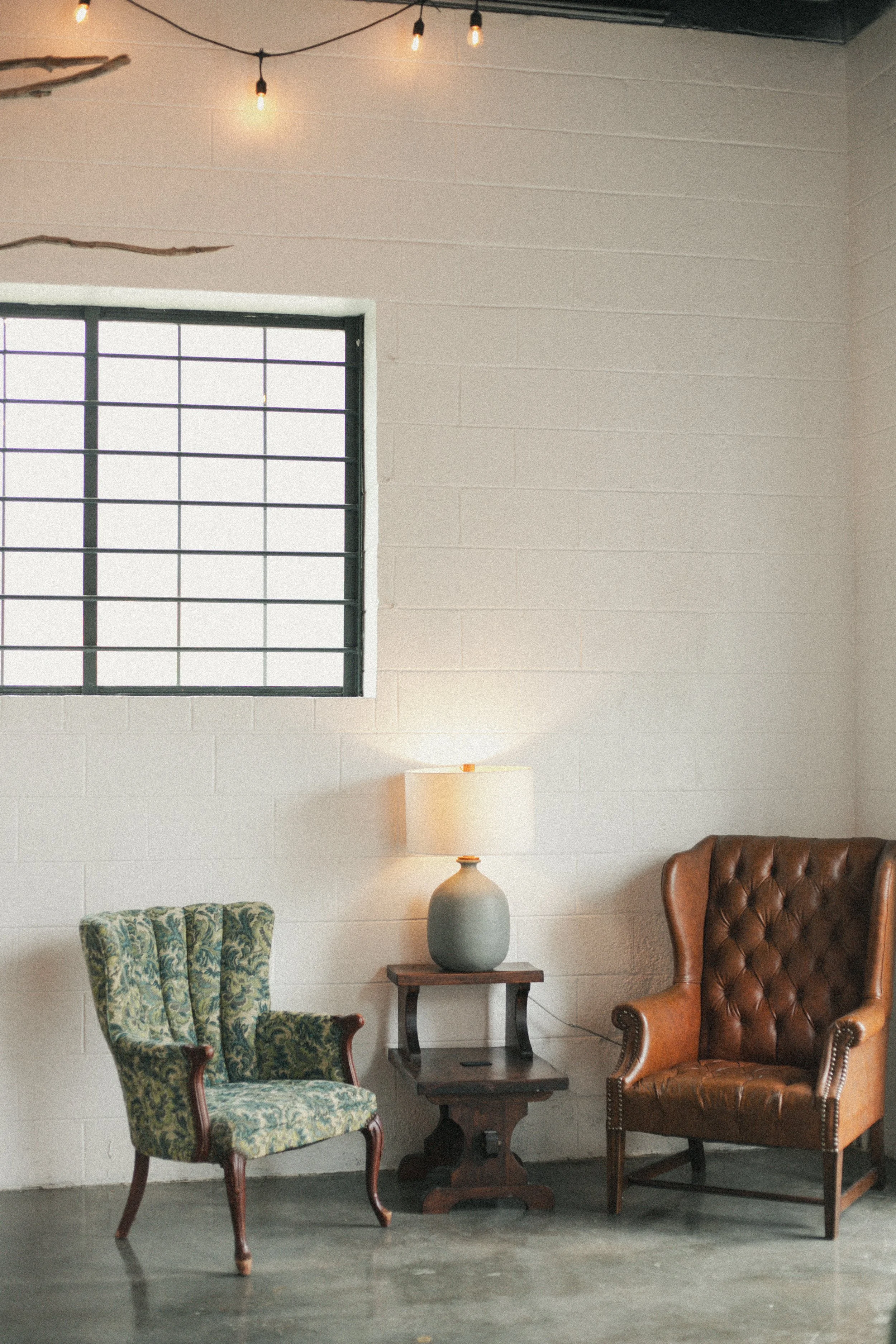 A cozy interior with vintage furniture, including a green patterned armchair, a brown leather armchair, a small wooden side table, and a table lamp with a white shade. There is a large window with black metal bars, white walls, and string lights hanging from the ceiling.