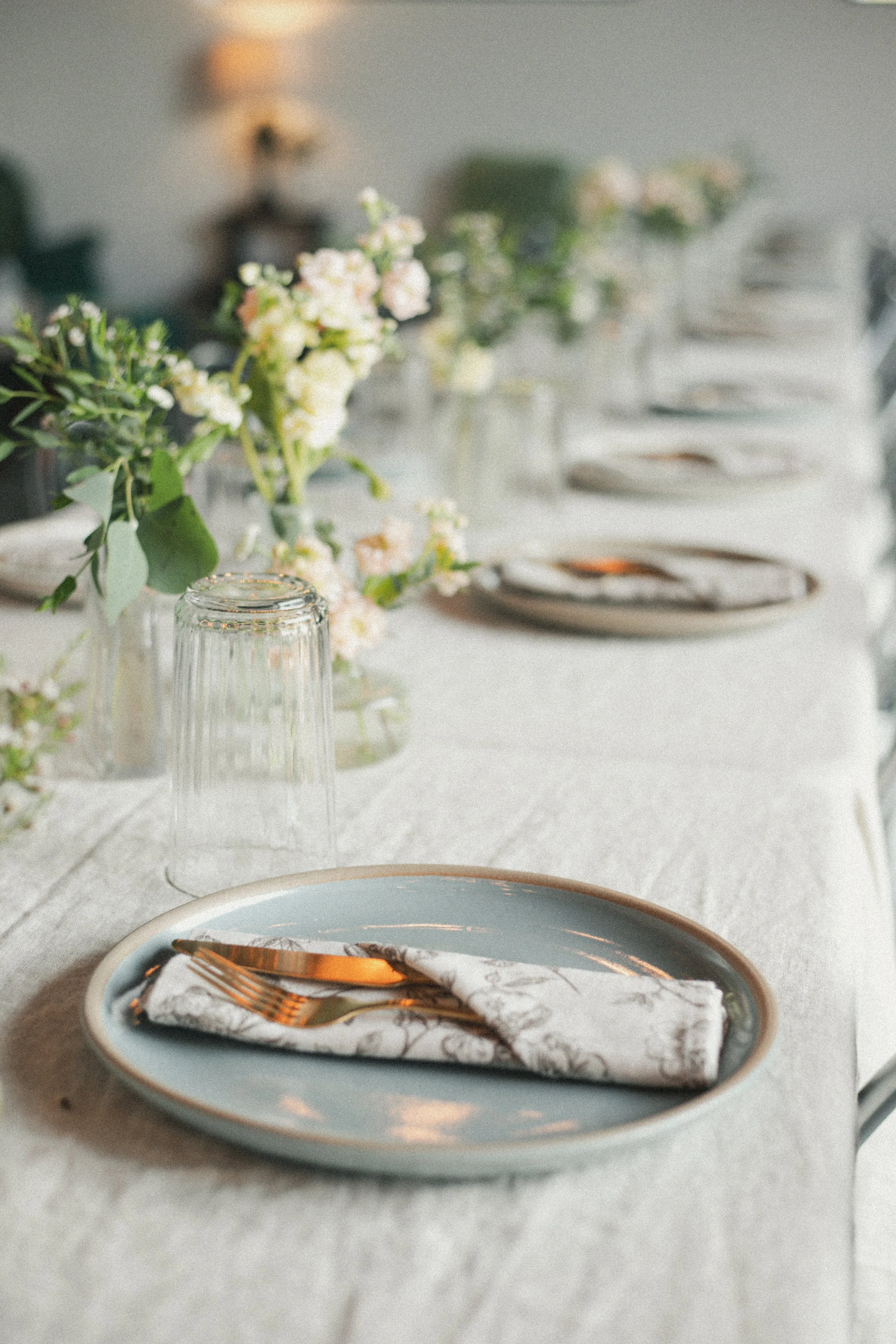 A table set with plates, forks, and napkins, with a centerpiece of white flowers and greenery, in a softly lit room.