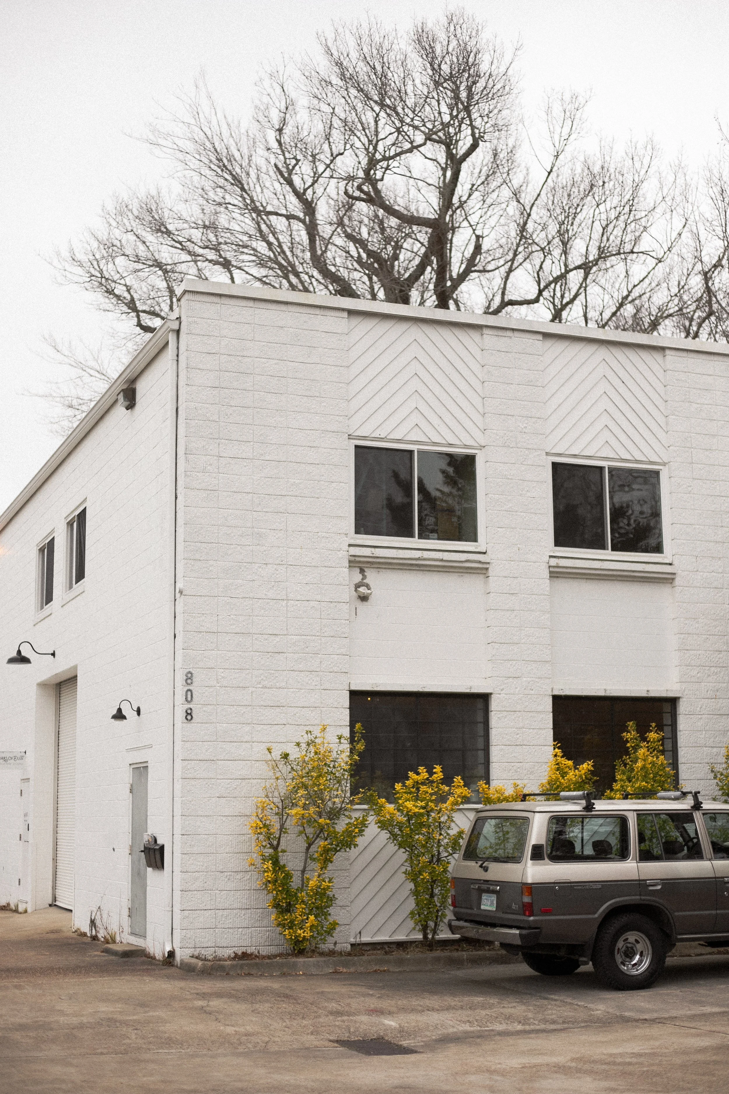 White modern two-story building with large windows and a garage, a gray SUV parked in front, and small trees with yellow flowers.